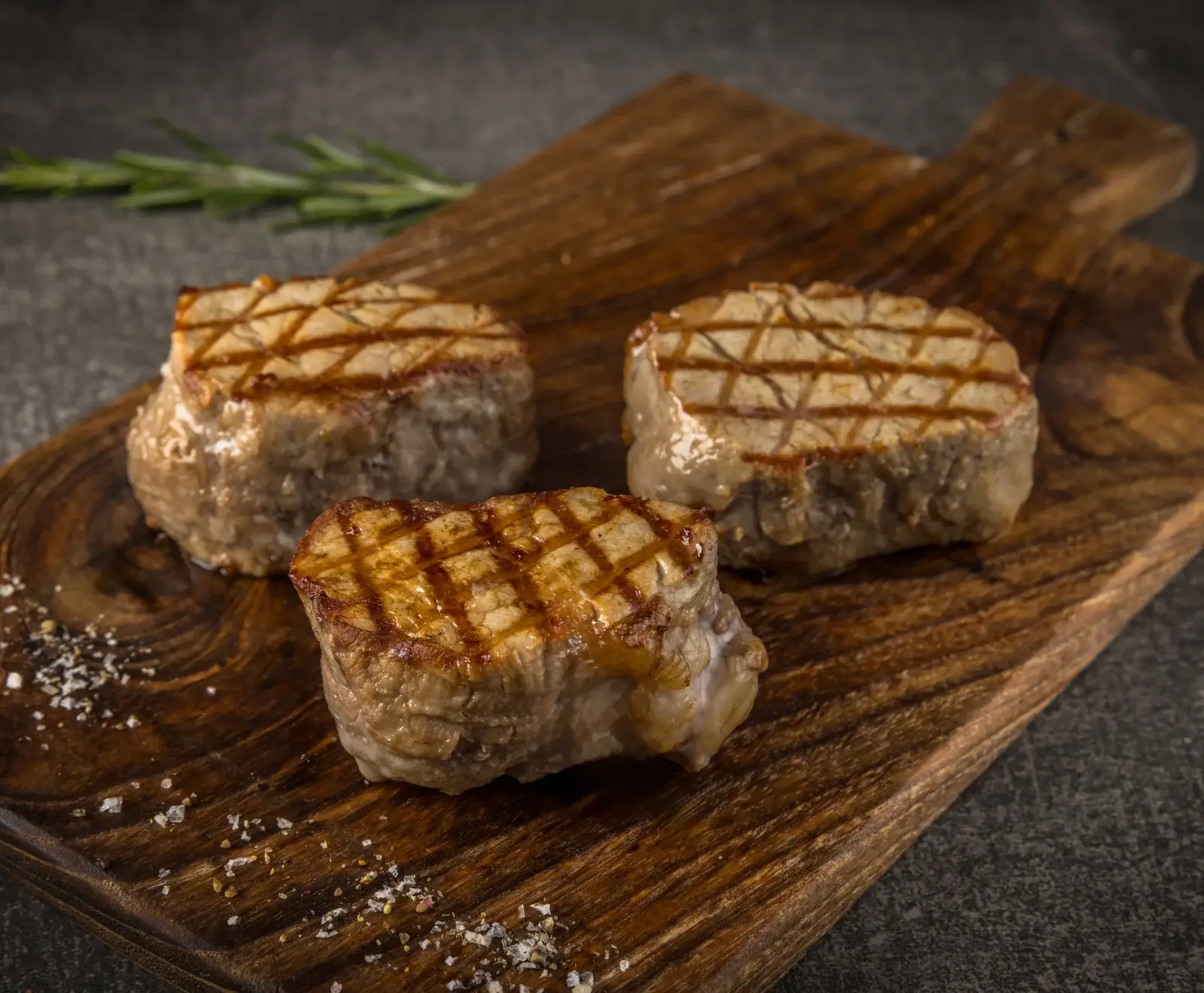 Drei saftige, gegrillte Fleischmedaillons mit deutlichen Grillstreifen liegen auf einem rustikalen Holzbrett, umgeben von grobem Salz und Pfeffer. Im Hintergrund ist ein Rosmarinzweig zu sehen.