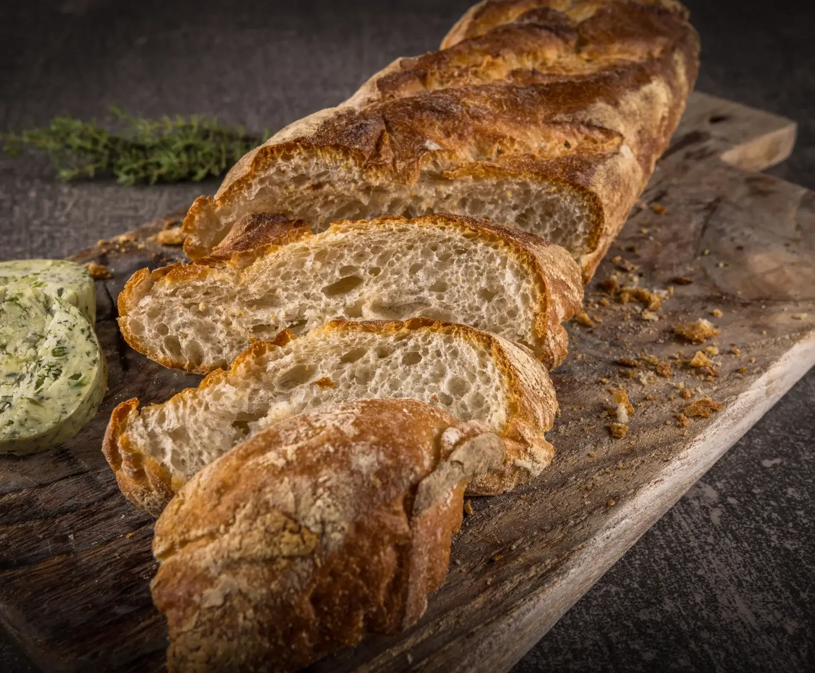 Ein angeschnittenes, knuspriges Baguette liegt auf einem rustikalen Holzbrett. Daneben sind zwei Scheiben Kräuterbutter und ein Thymianzweig zu sehen. Brotkrümel liegen auf dem Brett.