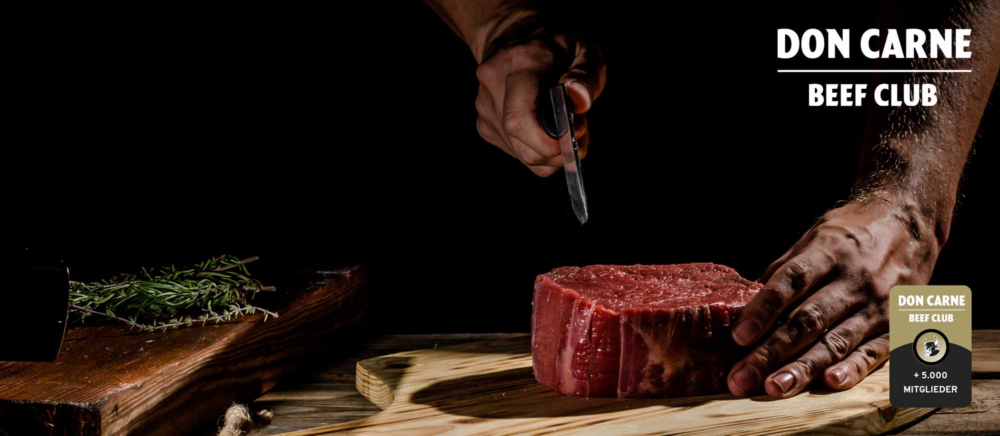 Hände würzen ein rohes, dickes Steak auf einem Holzbrett. Eine Hand hält ein Messer, die andere ruht auf dem Fleisch. Im Hintergrund Rosmarin. Logos: DON CARNE BEEF CLUB, + 5.000 MITGLIEDER.