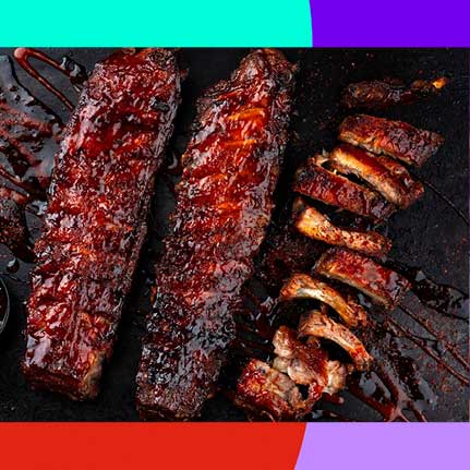Drei Rippchenstränge, zwei ganze und einer in einzelne Rippchen geschnitten, liegen auf einer dunklen Oberfläche und sind großzügig mit glänzender, dunkelroter Barbecue-Sauce bedeckt.
