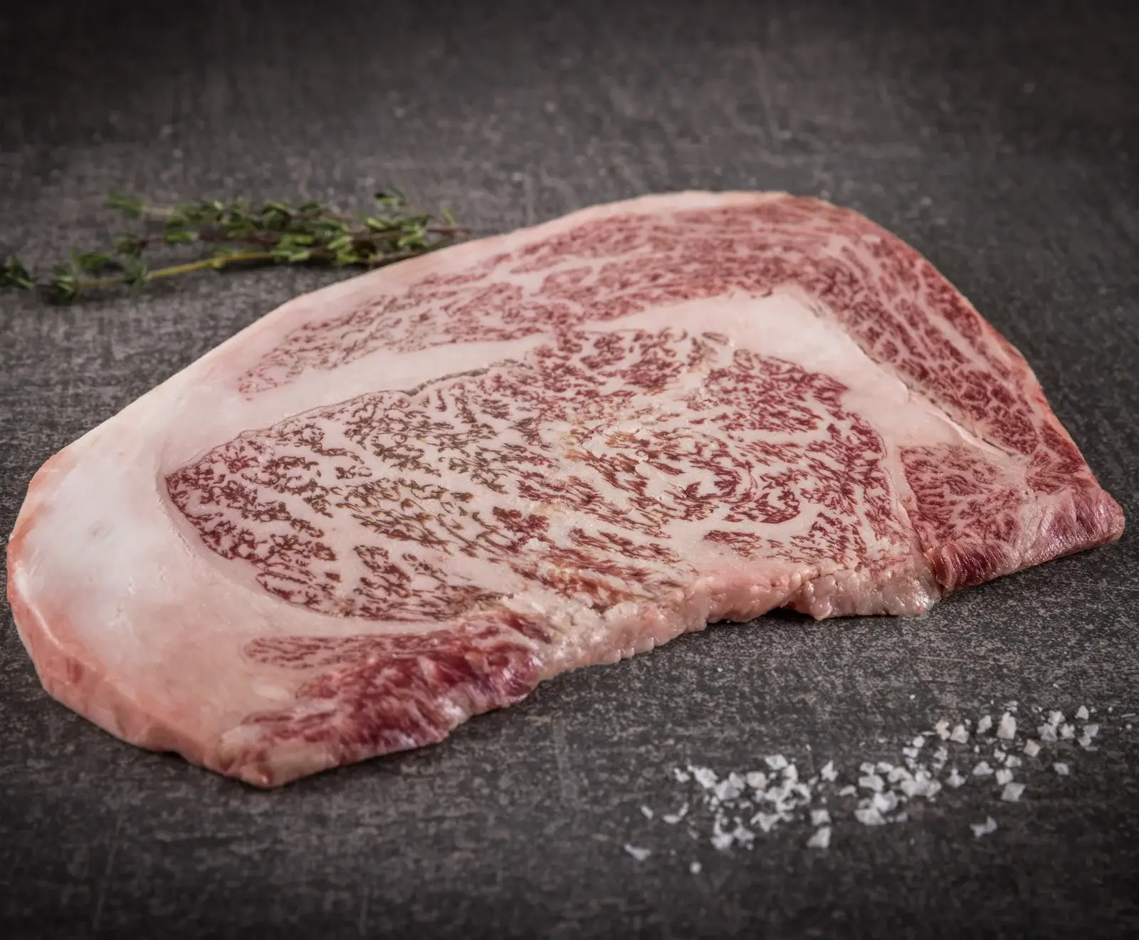 Ein rohes, stark marmoriertes Rindersteak (Wagyu-Stil) liegt auf einer dunklen Oberfläche, begleitet von einem Thymianzweig und Meersalzflocken.