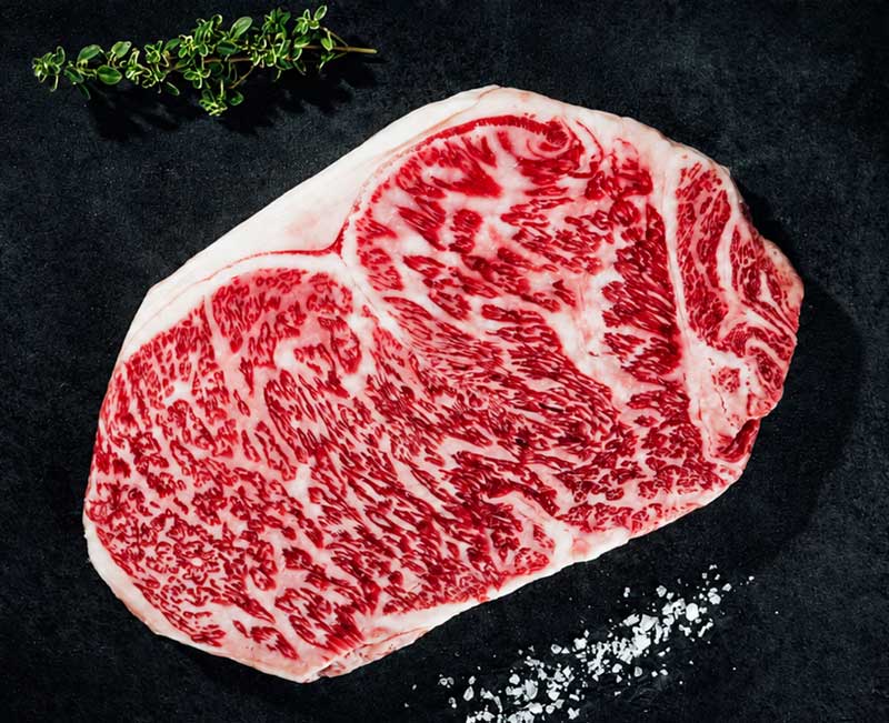 Ein rohes, stark marmoriertes Premium-Steak (z.B. Wagyu) liegt auf einer dunklen Oberfläche, begleitet von einem Thymianzweig oben links und grobem Salz unten rechts. Die intensive Fettmarmorierung ist deutlich sichtbar.