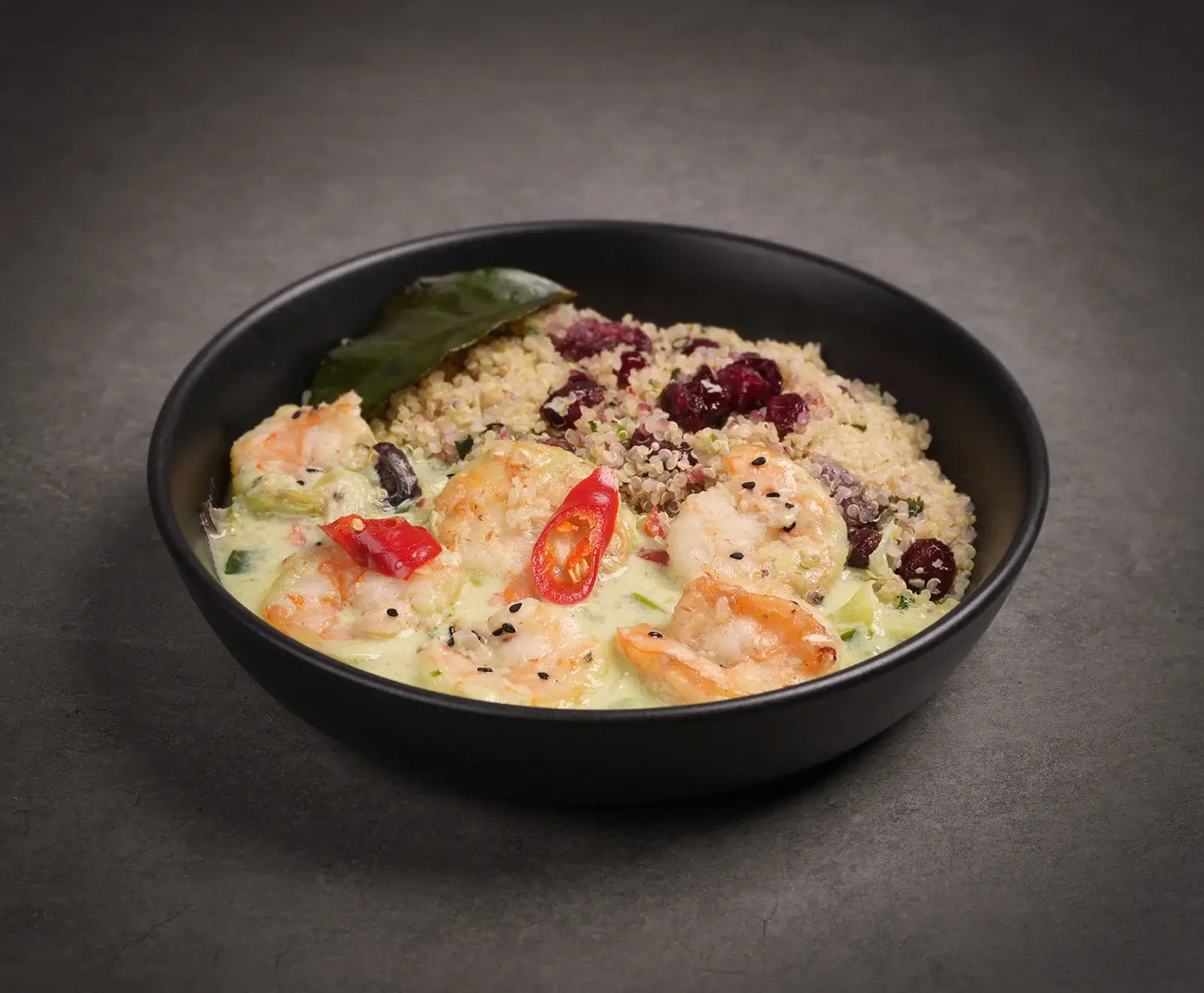 Schwarze Schale mit Garnelen in cremiger grüner Currysauce, garniert mit roten Chilischeiben und einem Blatt. Dazu Quinoa mit roten Beeren und schwarzem Sesam.