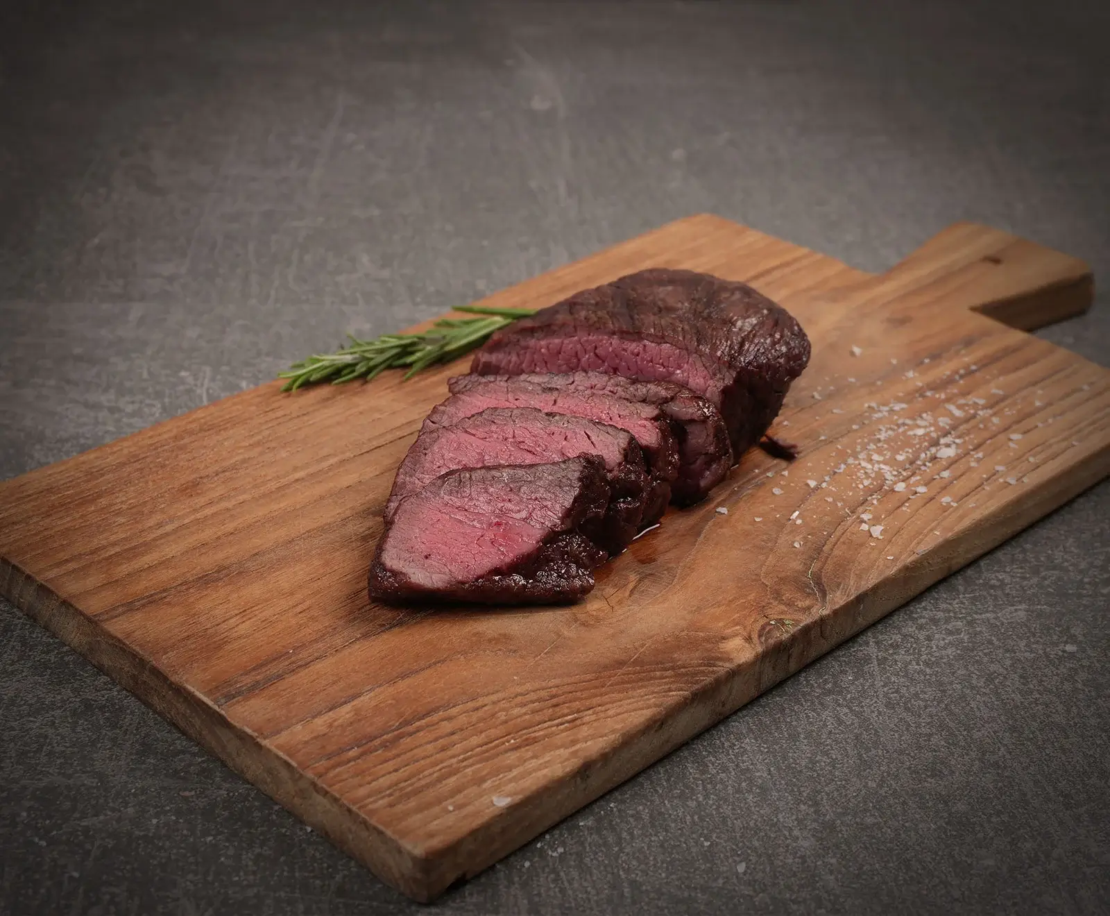 Ein aufgeschnittenes, medium-rare gebratenes Stück Fleisch auf einem Holzschneidebrett, garniert mit einem Rosmarinzweig und grobem Salz, auf dunklem Hintergrund.
