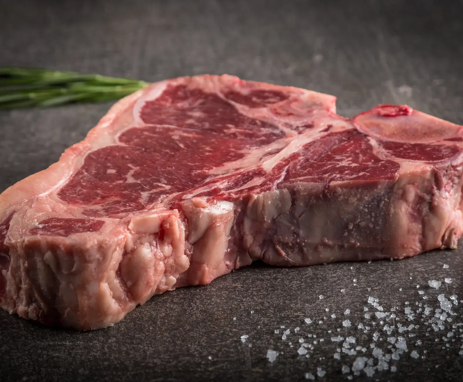 Rohes T-Bone-Steak mit deutlicher Marmorierung auf dunklem Untergrund, umgeben von grobem Salz und mit Rosmarin im unscharfen Hintergrund.