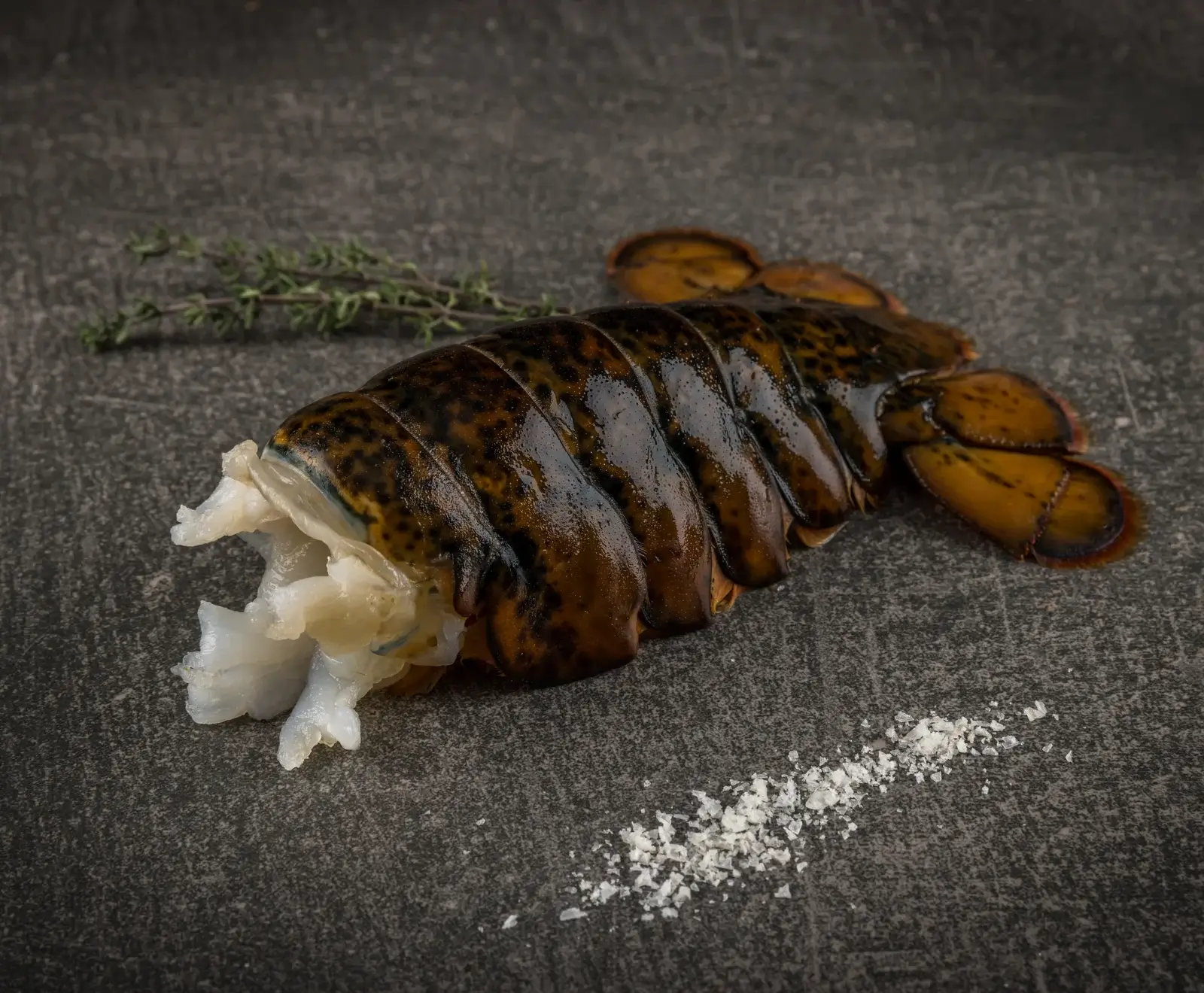 Roher Hummerschwanz, teilweise geschält mit sichtbarem weißen Fleisch, liegt auf dunklem Untergrund. Daneben liegen Thymianzweige und grobes Meersalz.