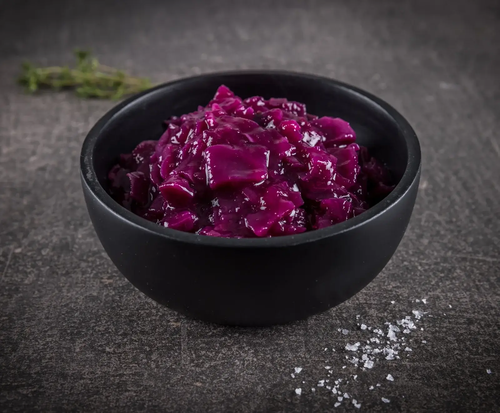Eine schwarze Schale, gefüllt mit leuchtend violettem, gekochtem Rotkohl, steht auf einem dunklen, strukturierten Untergrund. Im Hintergrund ist ein unscharfer Thymianzweig, im Vordergrund grobes Salz.