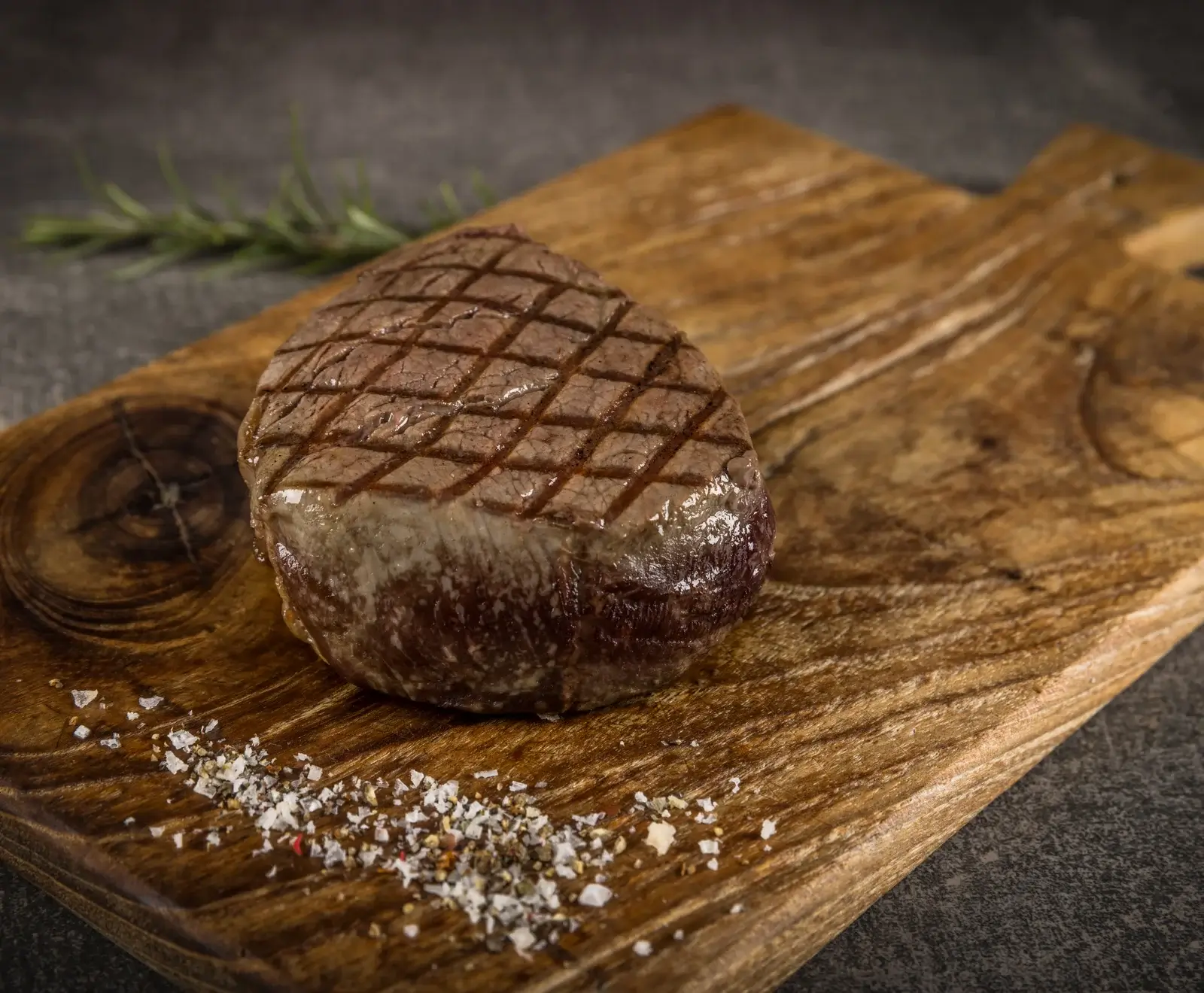 Ein perfekt gegrilltes Steak mit deutlichen Grillstreifen liegt auf einem rustikalen Holzbrett. Davor sind grobes Salz und Pfeffer verstreut, im Hintergrund ein Rosmarinzweig.
