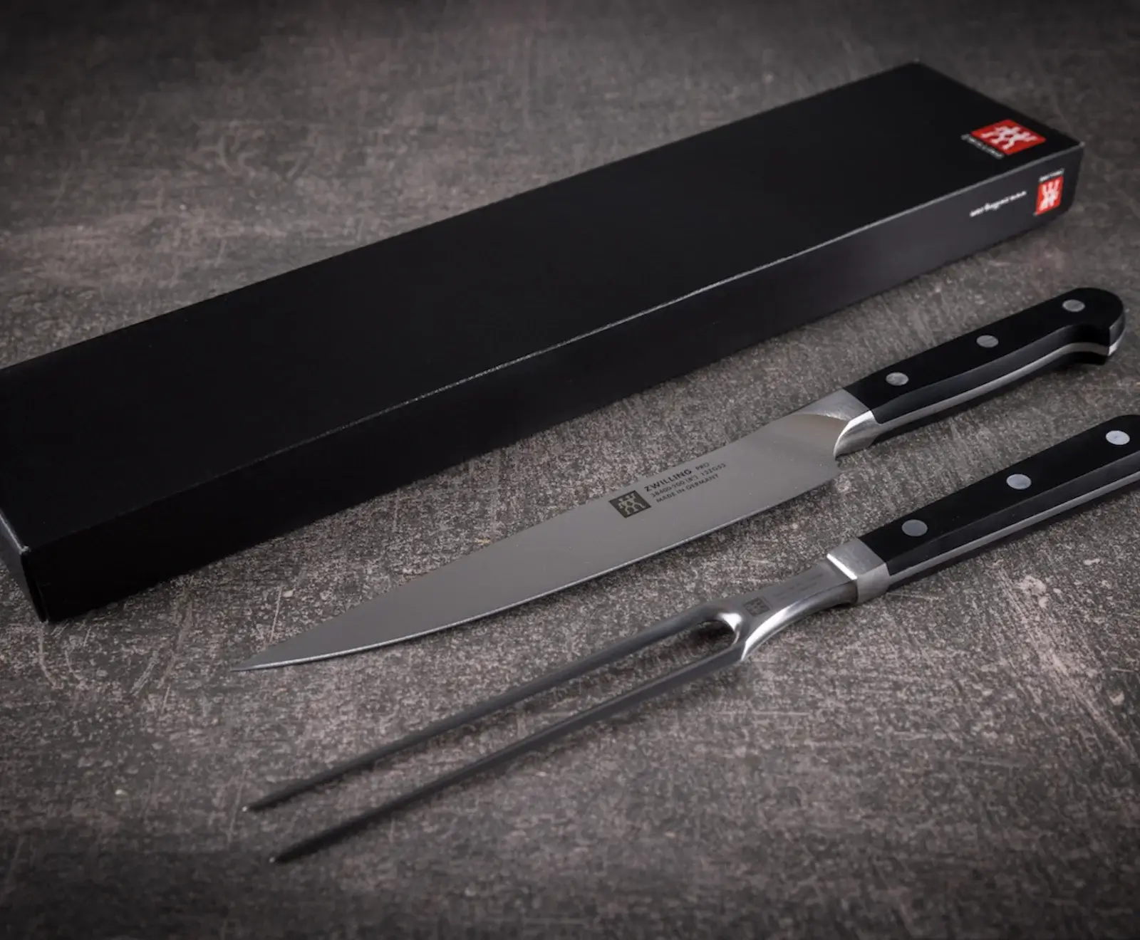 Ein Zwilling Pro Tranchierset, bestehend aus einem langen Messer und einer zweizinkigen Gabel mit schwarzen Griffen, liegt auf einer dunklen Oberfläche. Im Hintergrund ist die schwarze Verpackung mit dem Zwilling-Logo zu sehen.