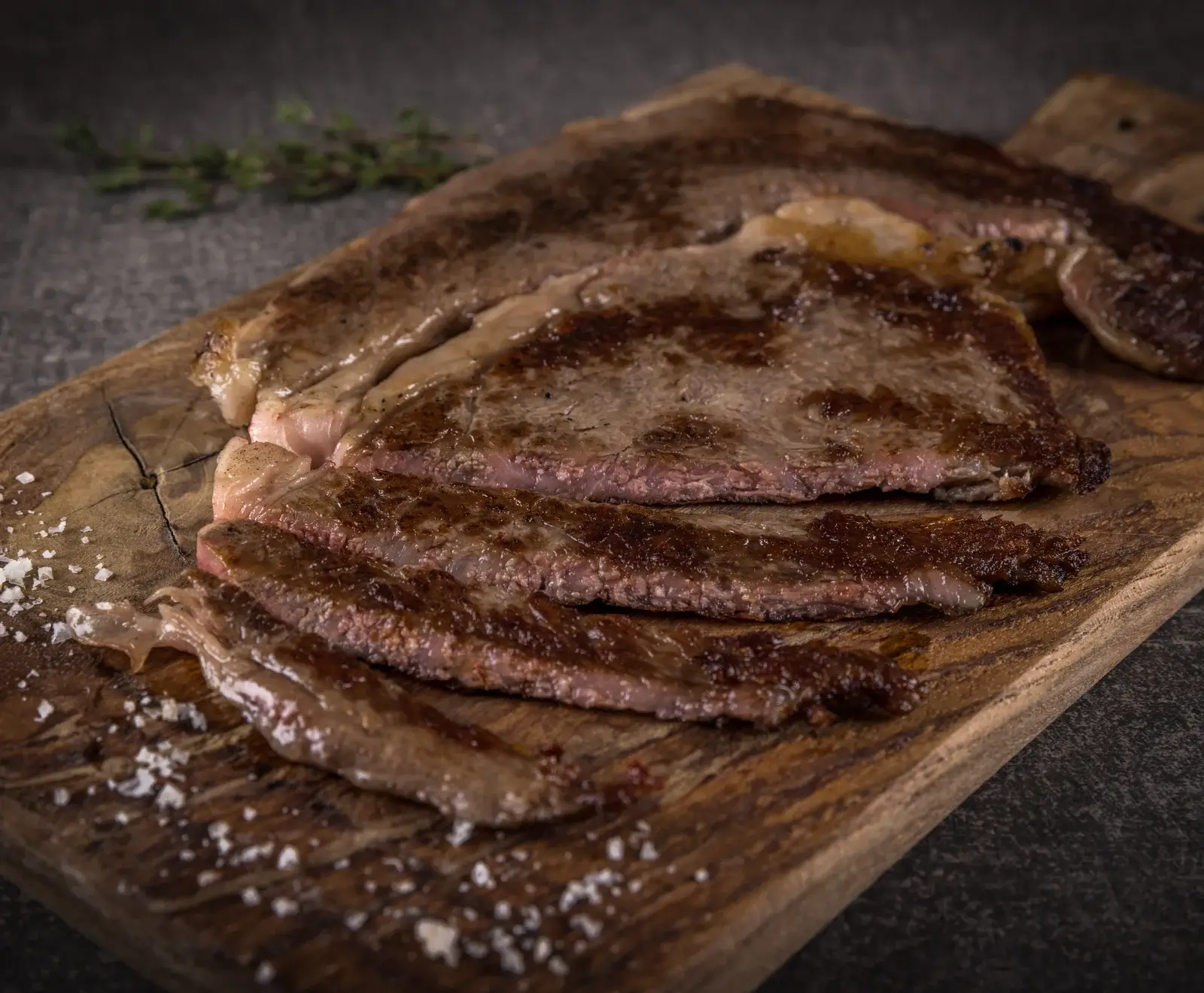 Aufgeschnittenes, medium gebratenes Steak auf einem rustikalen Holzbrett, bestreut mit grobem Meersalz. Im Hintergrund ein unscharfer Kräuterzweig.