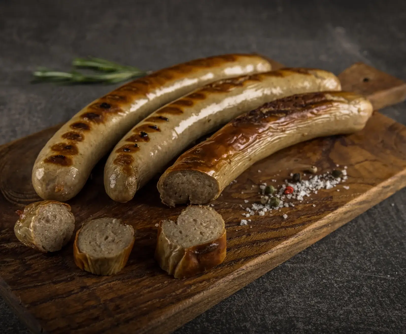 Drei goldbraun gegrillte Bratwürste auf einem rustikalen Holzbrett. Eine Wurst ist angeschnitten, daneben liegen grobes Salz, bunte Pfefferkörner und Rosmarin. Der Hintergrund ist dunkel und strukturiert.