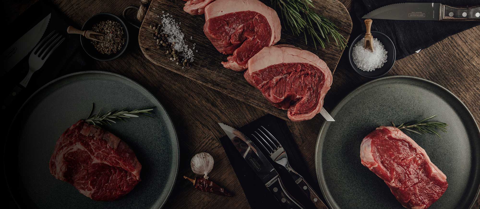 Nahaufnahme von rohem Rindfleisch auf einem Holztisch, umgeben von Rosmarin, Salz, Pfeffer, Knoblauch und Besteck. Zwei Steaks auf einem Brett, zwei auf dunklen Tellern.