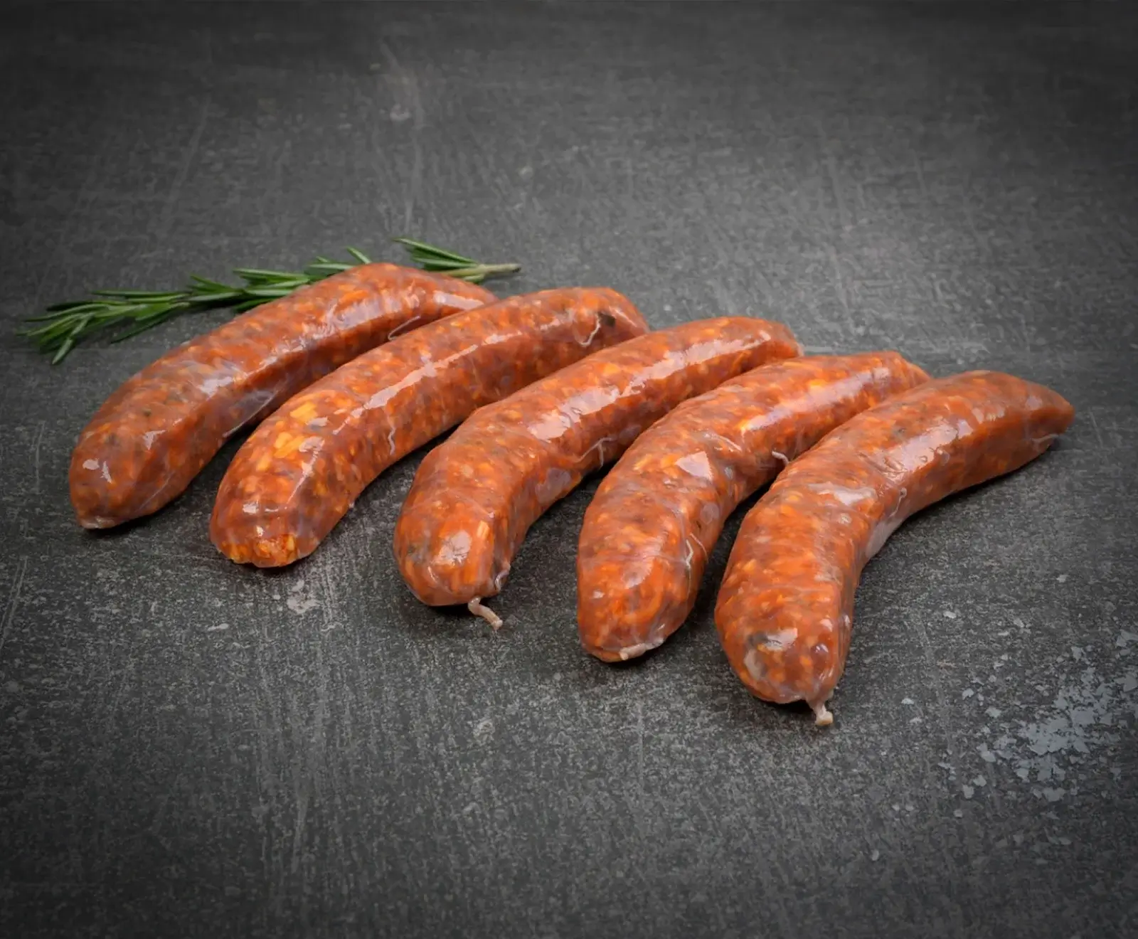 Sechs rohe, rötlich-orangefarbene Würste liegen leicht gekrümmt auf einer dunklen, strukturierten Oberfläche. Links oben ist ein Rosmarinzweig zu sehen. Die Würste wirken frisch und sind bereit zur Zubereitung.