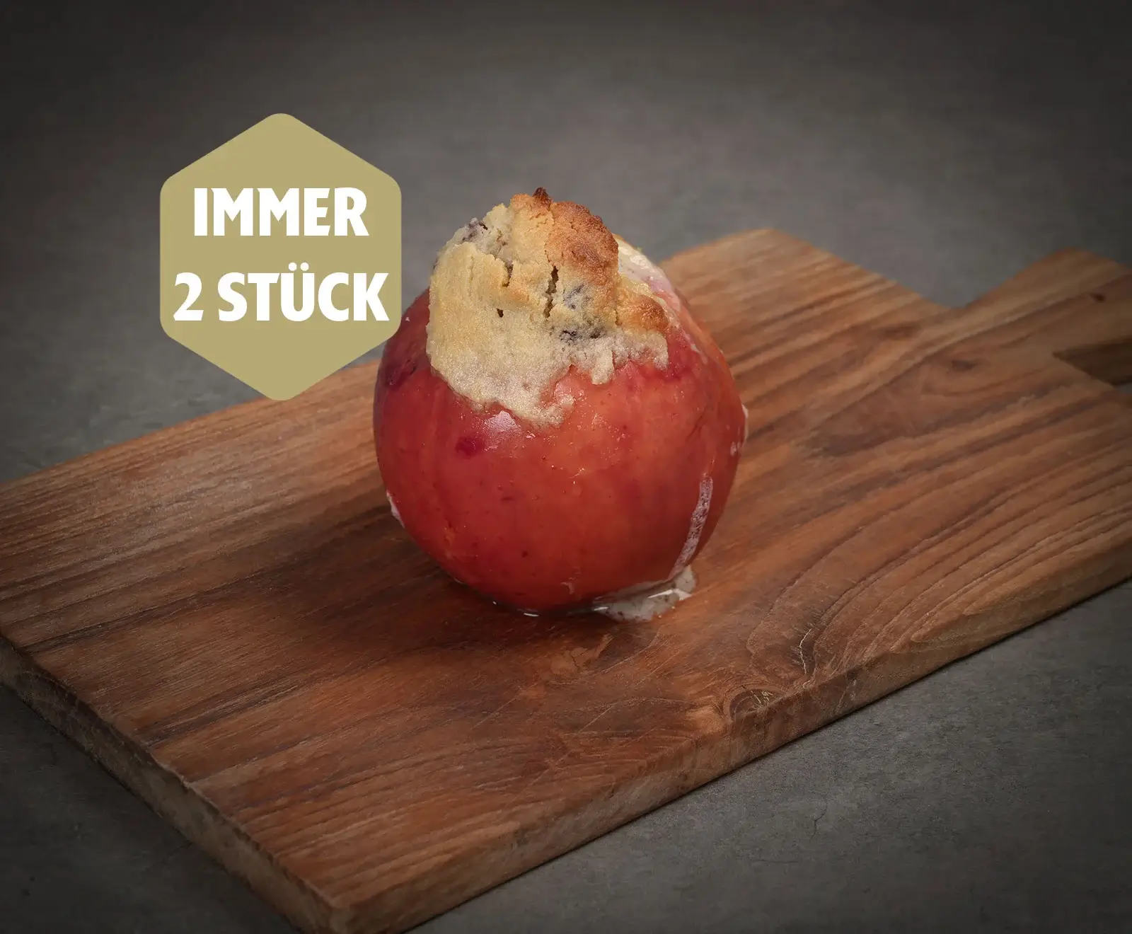 Ein saftiger, rot gebackener Apfel mit goldbraunen Streuseln auf einem Holzbrett. Etwas Füllung ist ausgelaufen. Oben links ein goldenes Schild mit der Aufschrift "IMMER 2 STÜCK".
