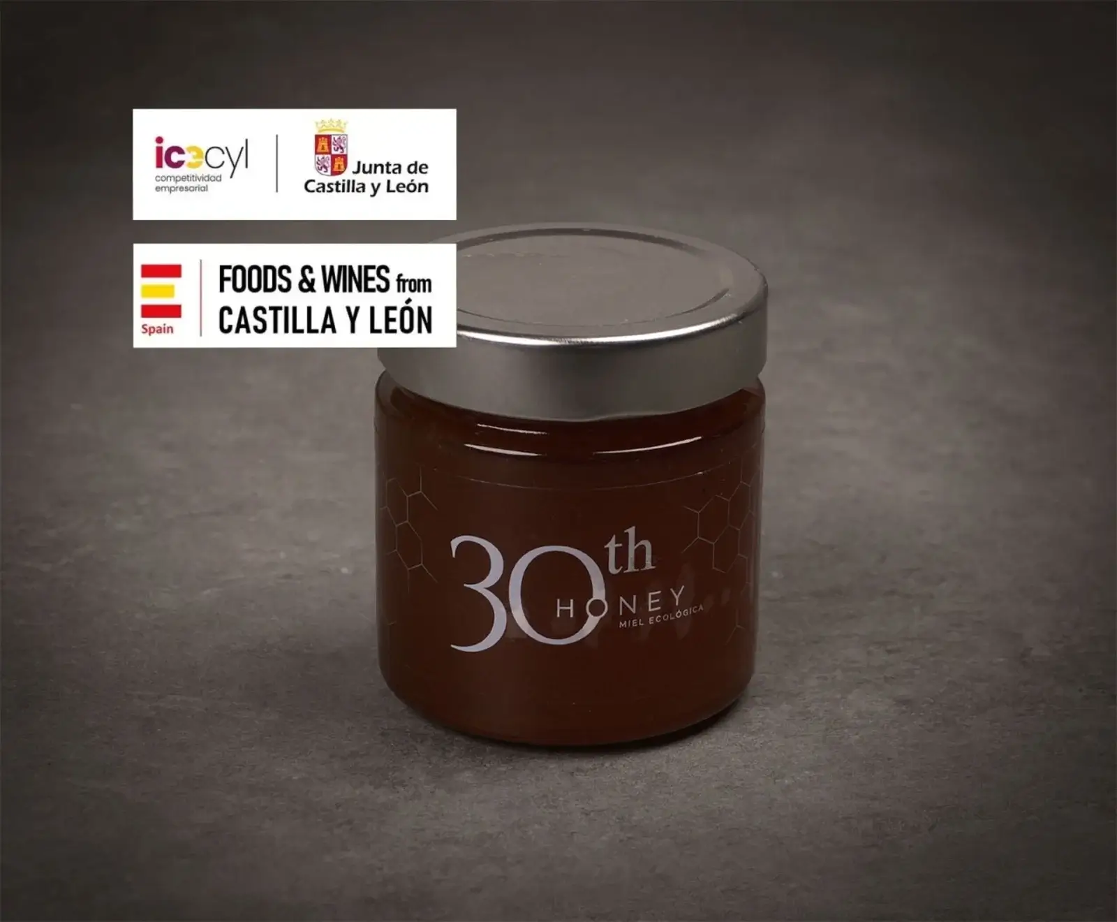 Ein Glas Honig mit silbernem Deckel und der Aufschrift "30th HONEY Miel Ecológica" steht auf dunklem Untergrund. Oben links sind Logos von "icecyl", "Junta de Castilla y León" und "FOODS & WINES from CASTILLA Y LEÓN" zu sehen.