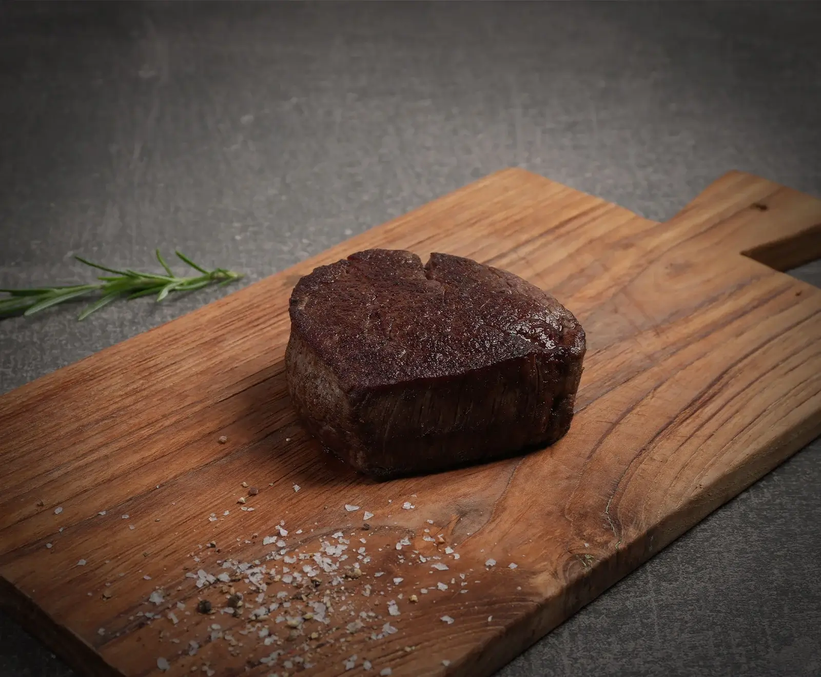 Ein perfekt gebratenes, dickes Stück Rinderfilet liegt auf einem rustikalen Holzschneidebrett. Grobe Salzkristalle und Pfeffer sind daneben verstreut, ein Rosmarinzweig liegt im Hintergrund.