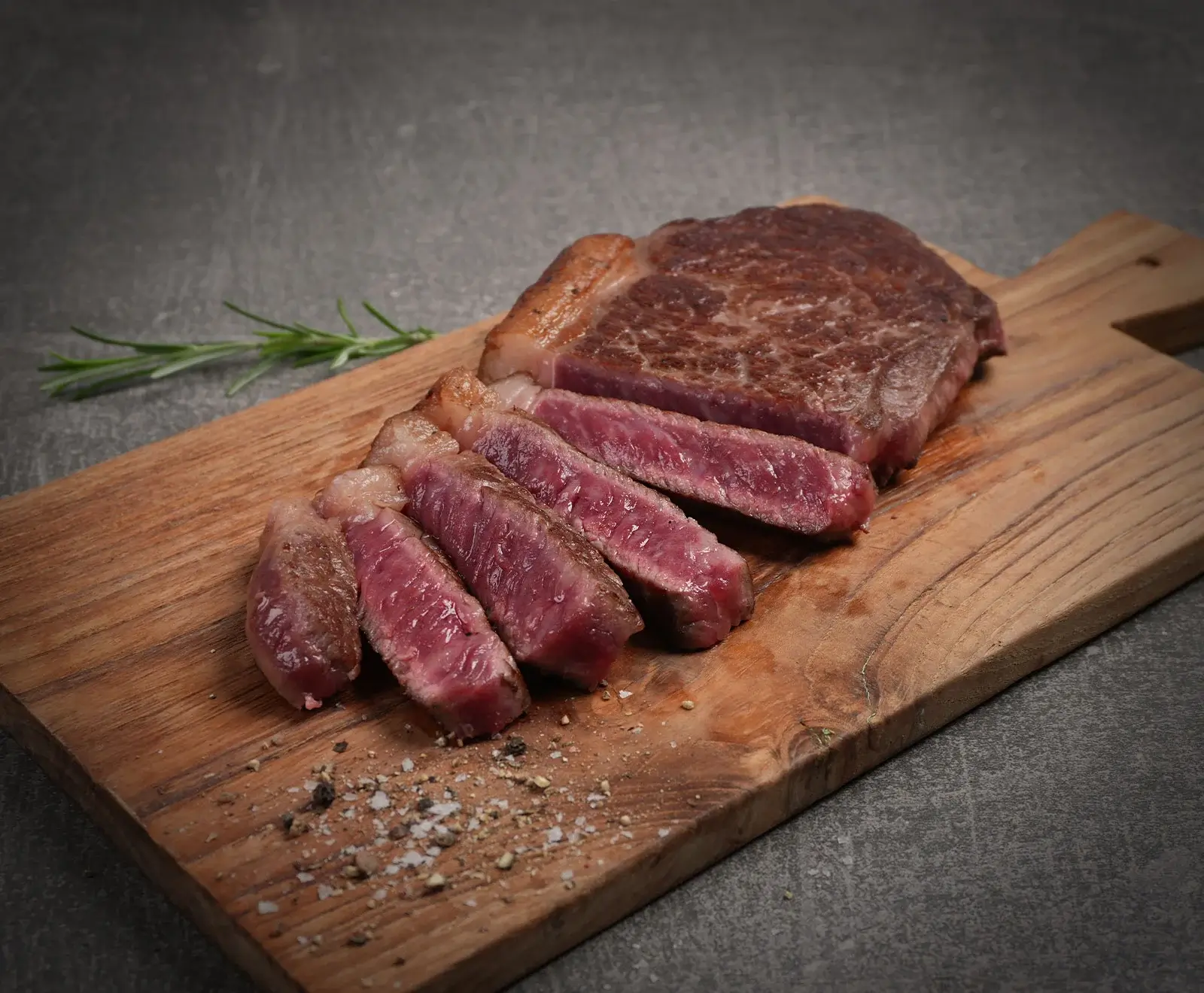 Ein medium-rare gebratenes Rindersteak, teilweise in Scheiben geschnitten, auf einem Holzschneidebrett. Grobes Salz, Pfeffer und ein Rosmarinzweig sind sichtbar.