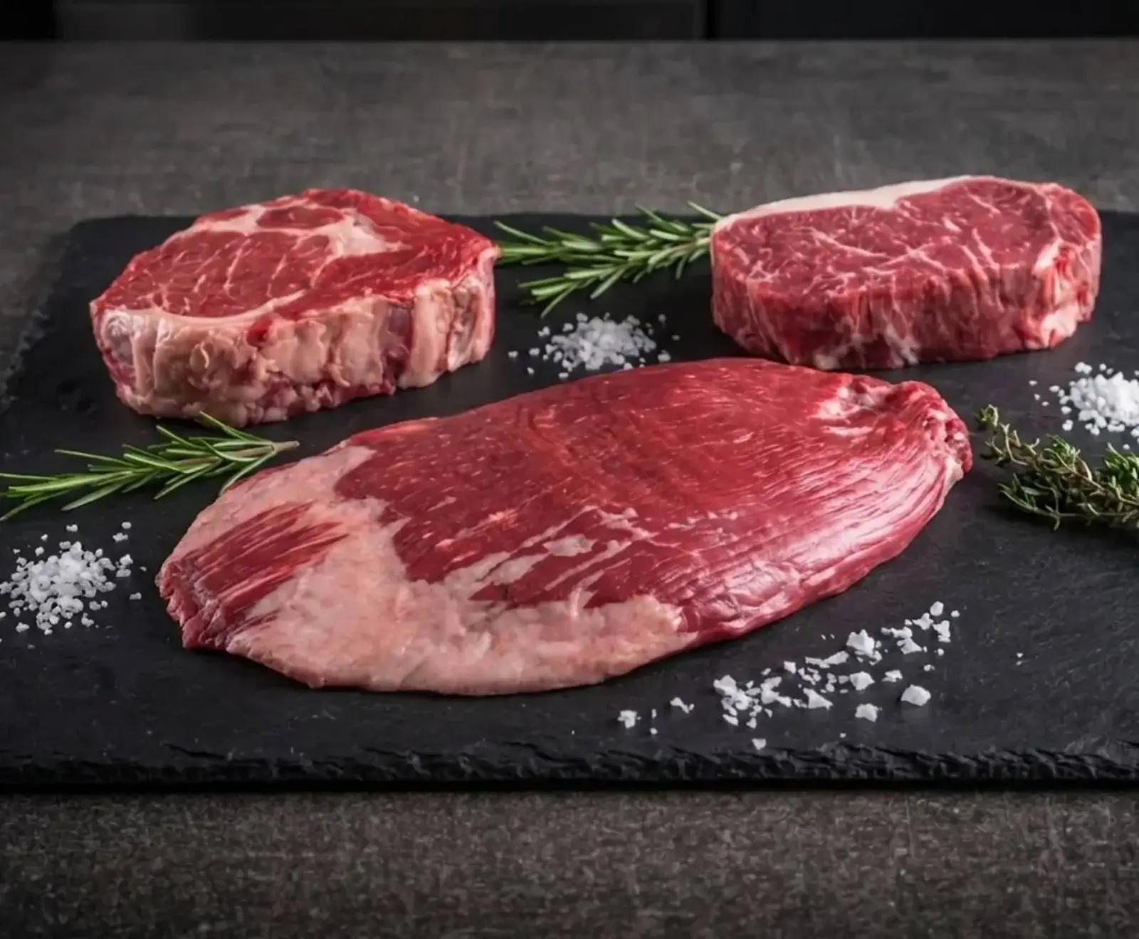 Drei rohe Fleischstücke (Ribeye, Roastbeef, Flanksteak) liegen auf einer dunklen Schieferplatte, umgeben von Rosmarin, Thymian und grobem Meersalz.