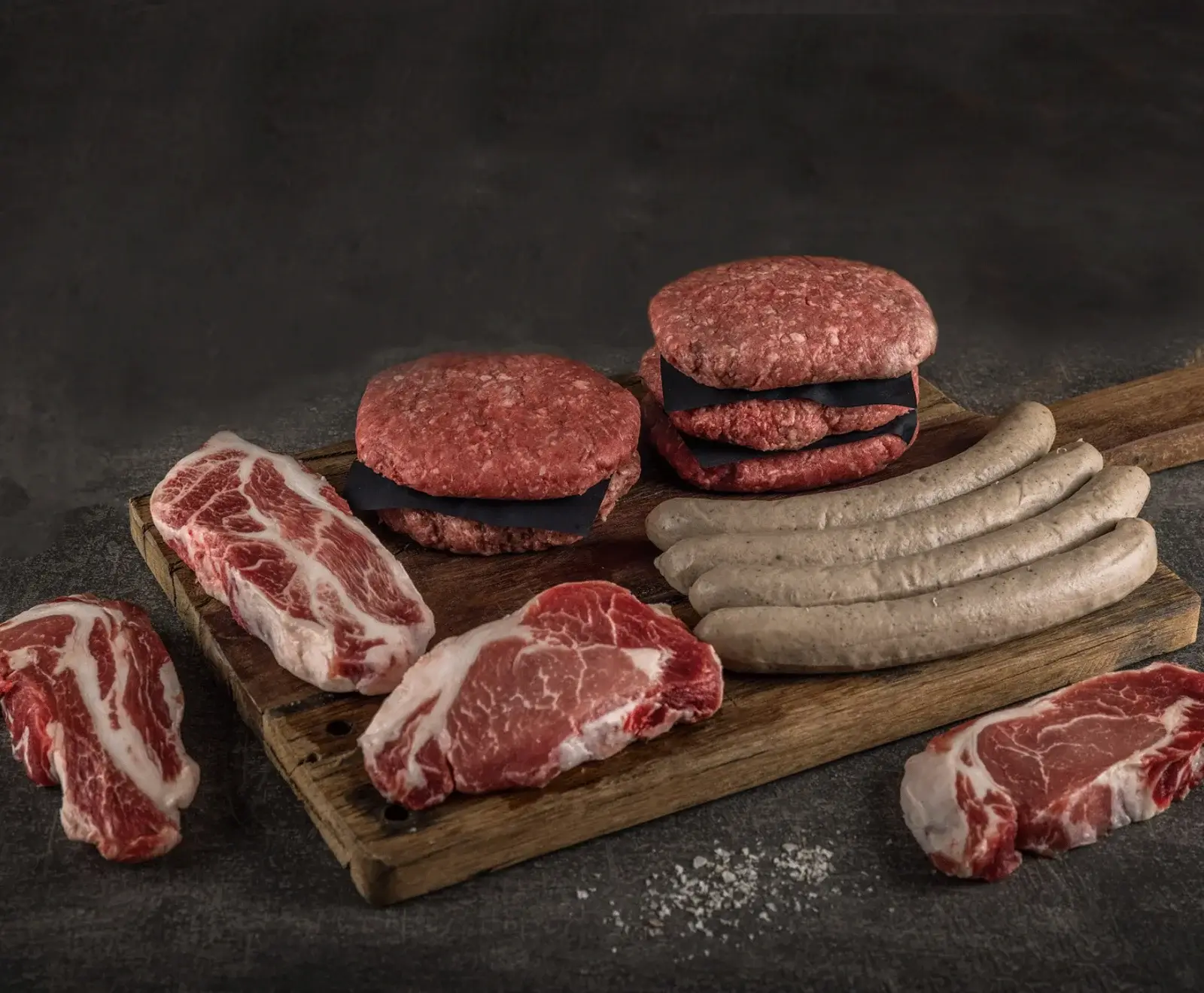 Verschiedene rohe Fleischprodukte auf einem Holzbrett: Burger-Patties mit Trennpapier, helle Würste und marmorierte Steaks, auf dunklem Hintergrund mit Salz.
