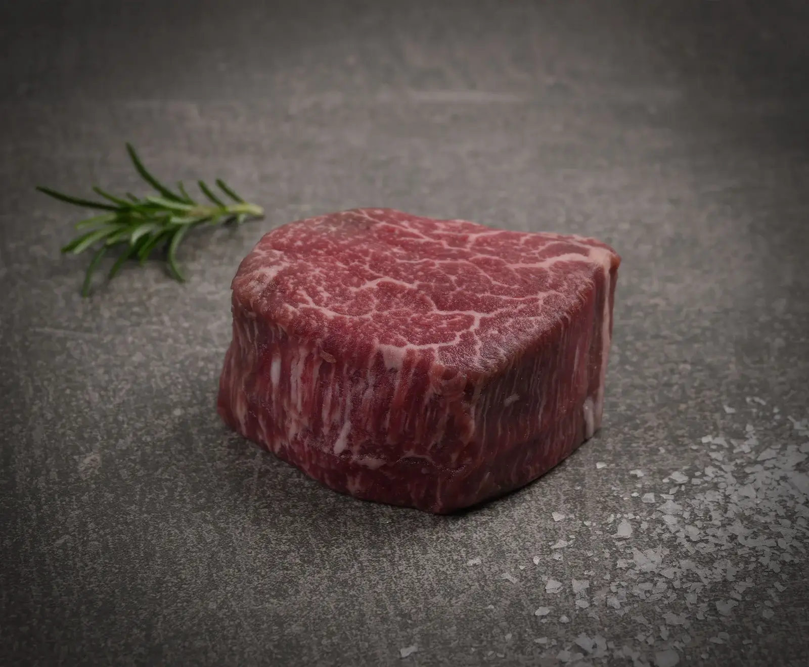 Ein rohes, stark marmoriertes Rinderfiletsteak liegt auf einem dunklen, strukturierten Untergrund, begleitet von einem Rosmarinzweig und groben Meersalzflocken.