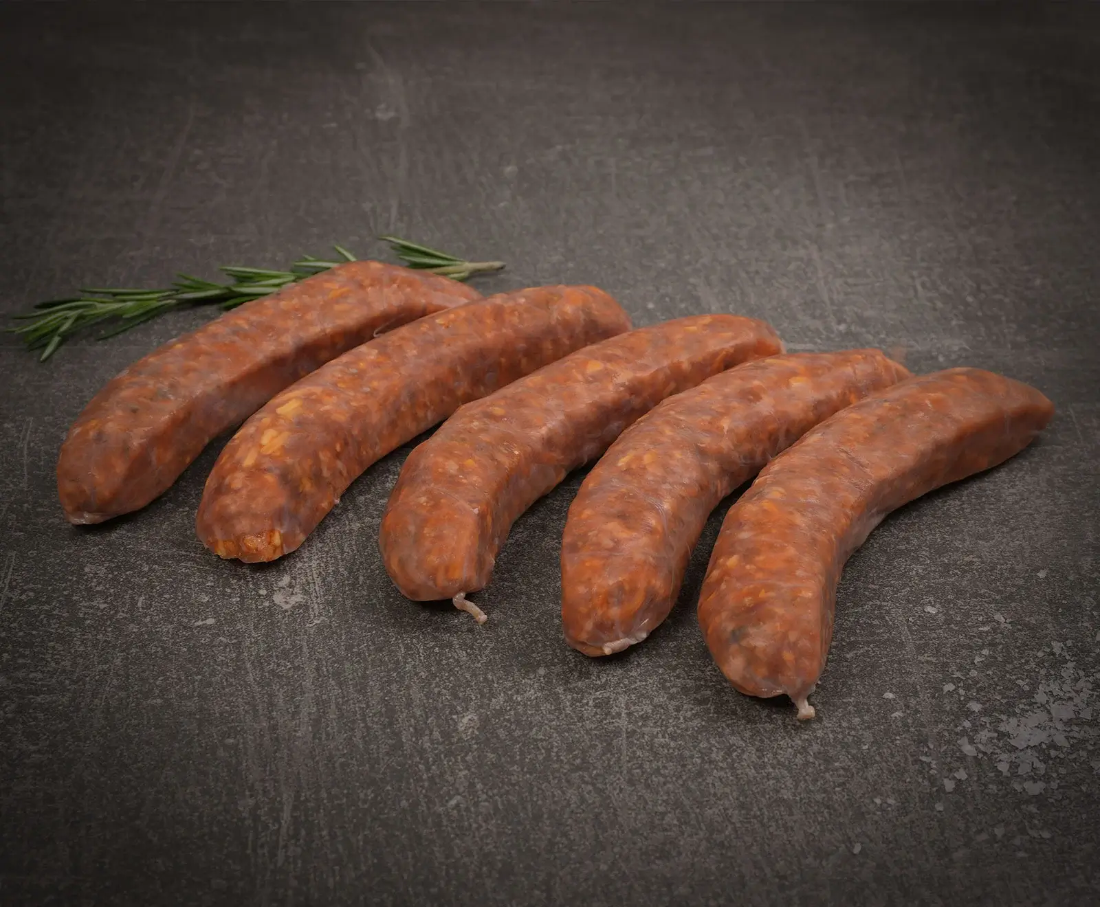 Fünf rohe, rötlich-braune Würste liegen diagonal auf einer dunklen, strukturierten Oberfläche. Im Hintergrund links sind einige Rosmarinzweige zu sehen.