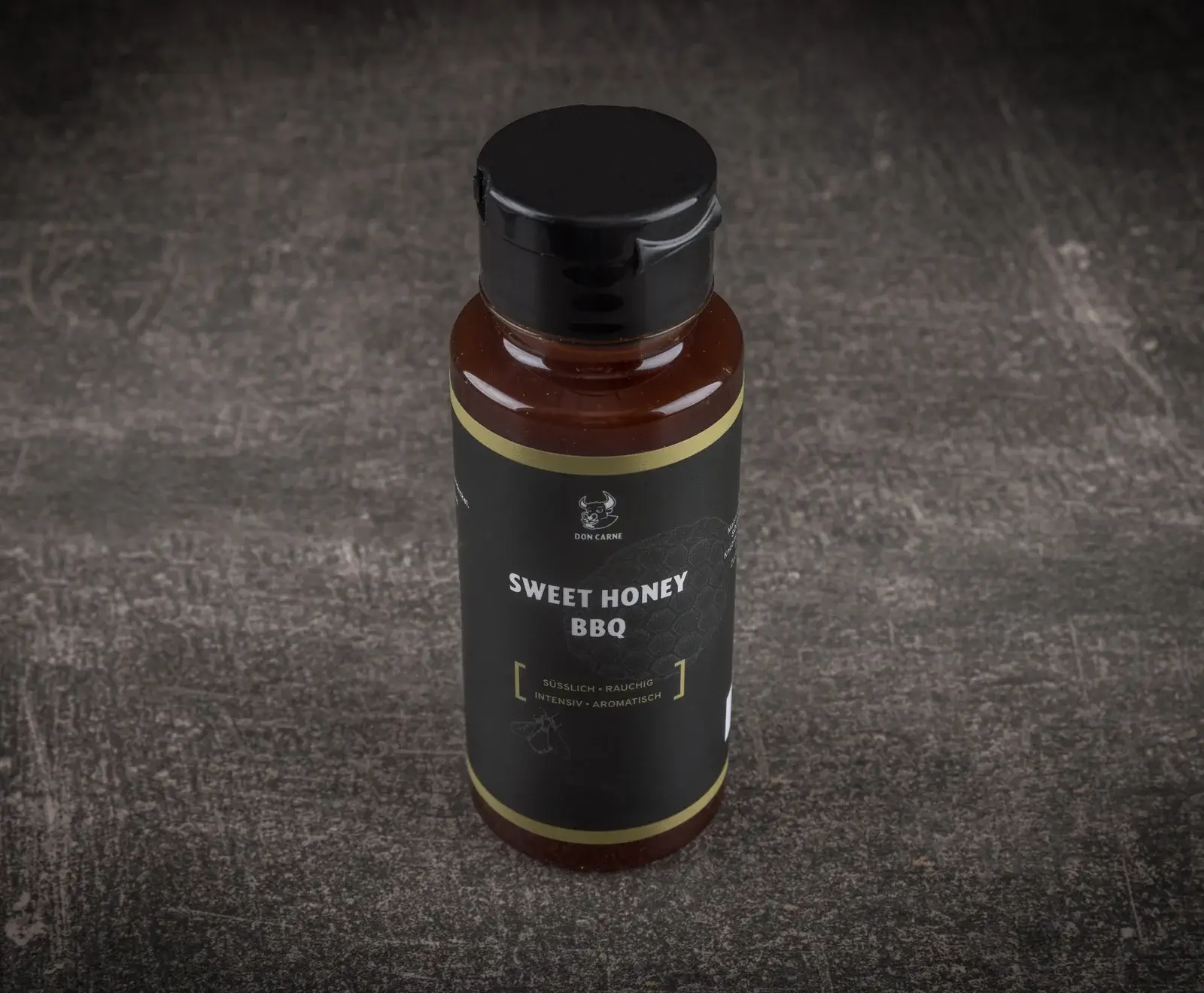 Eine Flasche Don Carne Sweet Honey BBQ Sauce mit schwarzem Etikett, goldener Umrandung und schwarzem Klappdeckel, stehend auf einer dunklen, strukturierten Oberfläche.