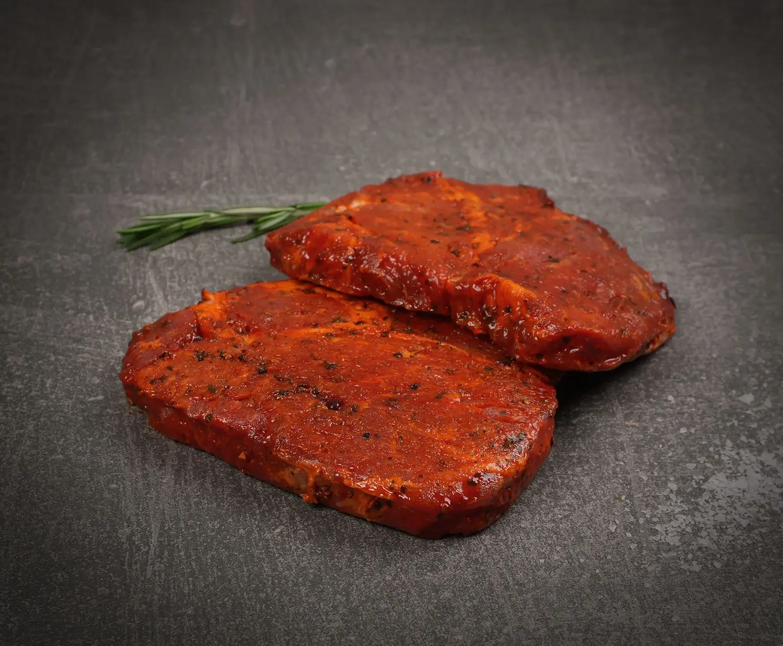 Zwei rohe, rot marinierte Fleischsteaks liegen auf einem dunklen, strukturierten Untergrund. Die Steaks sind mit Kräutern und Gewürzen bestreut. Im Hintergrund ist ein Rosmarinzweig zu sehen.