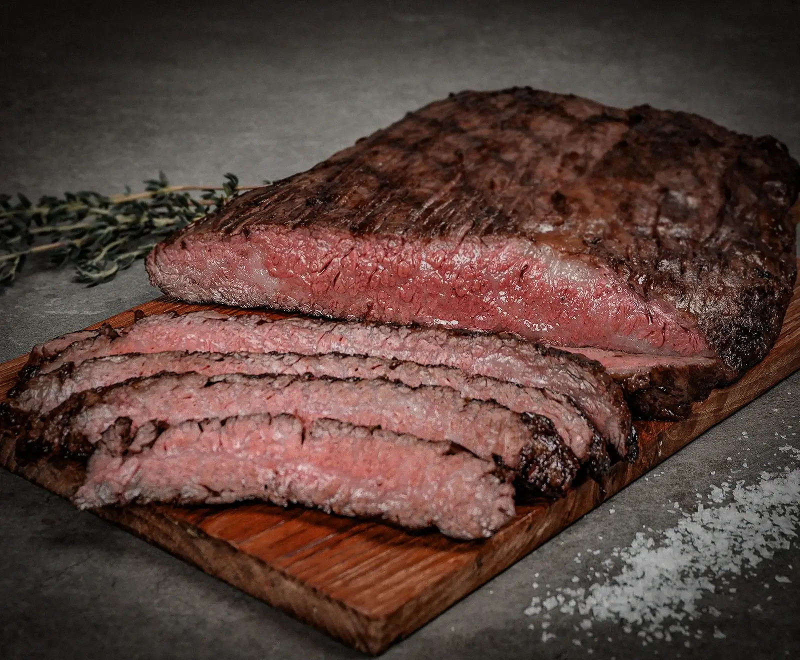 Ein saftiges, medium-rare gegartes Stück Fleisch, teilweise in Scheiben geschnitten, liegt auf einem Holzbrett. Im Hintergrund sind Thymianzweige und grobes Salz zu sehen.