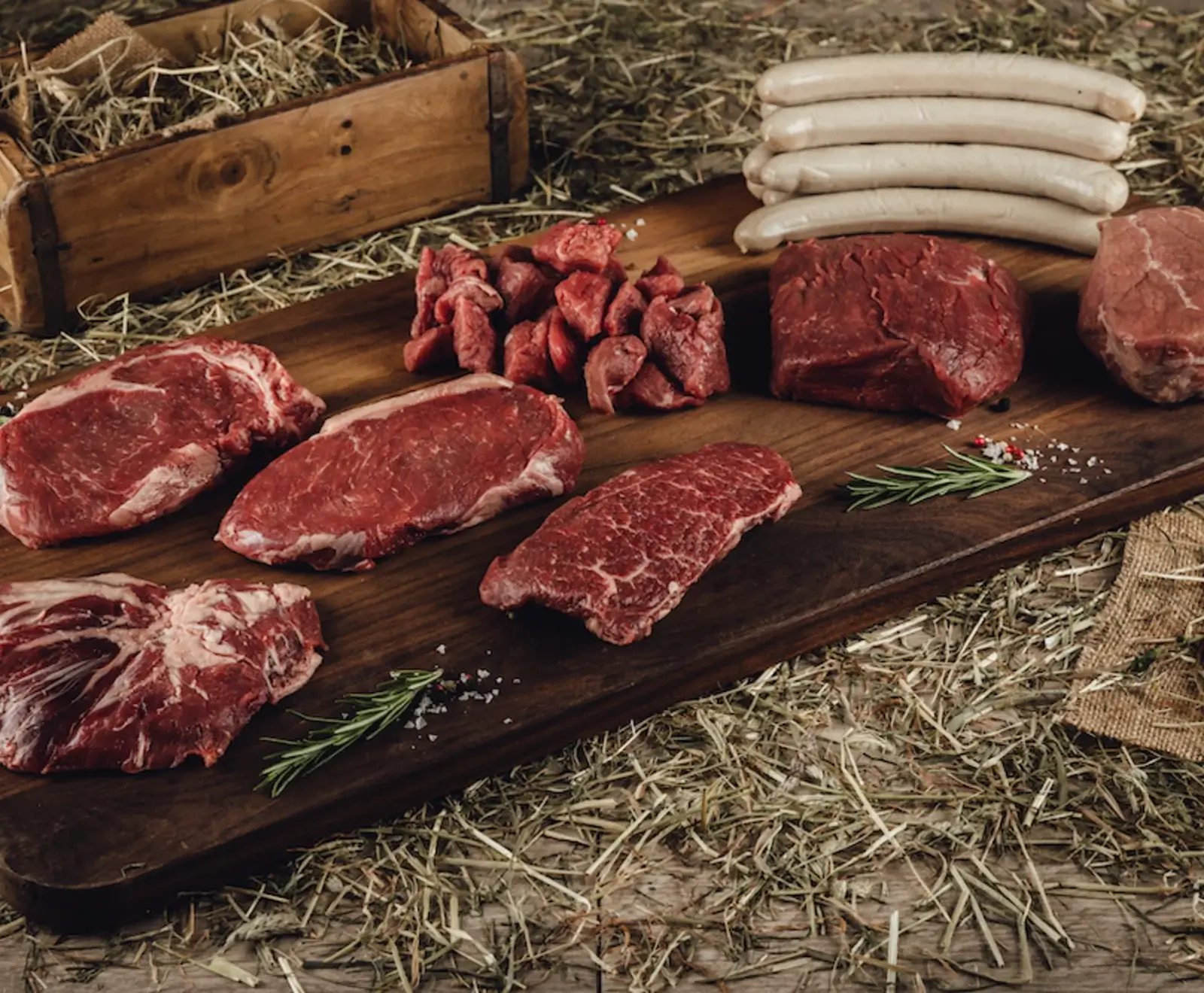 Rohfleisch-Auswahl auf Holzbrett: Steaks, Würfel, Bratenstücke und weiße Würste, garniert mit Rosmarin und Gewürzen, auf Heu und Holztisch.