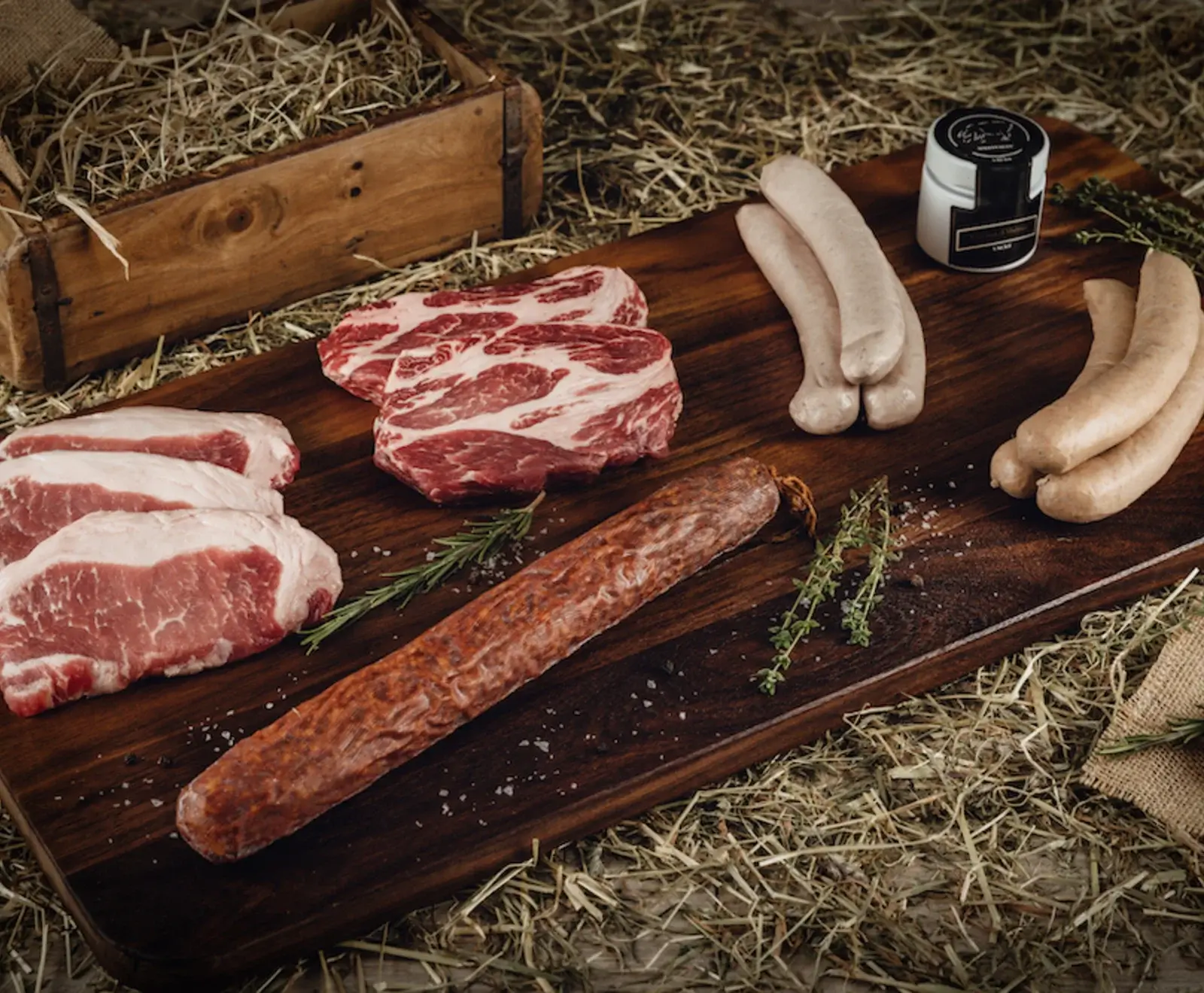 Eine rustikale Holzplatte mit verschiedenen rohen Fleisch- und Wurstwaren: drei Schweinekoteletts, zwei Rindersteaks, eine Salami, sechs helle Würste, garniert mit Rosmarin, Thymian und Meersalz auf Heu.
