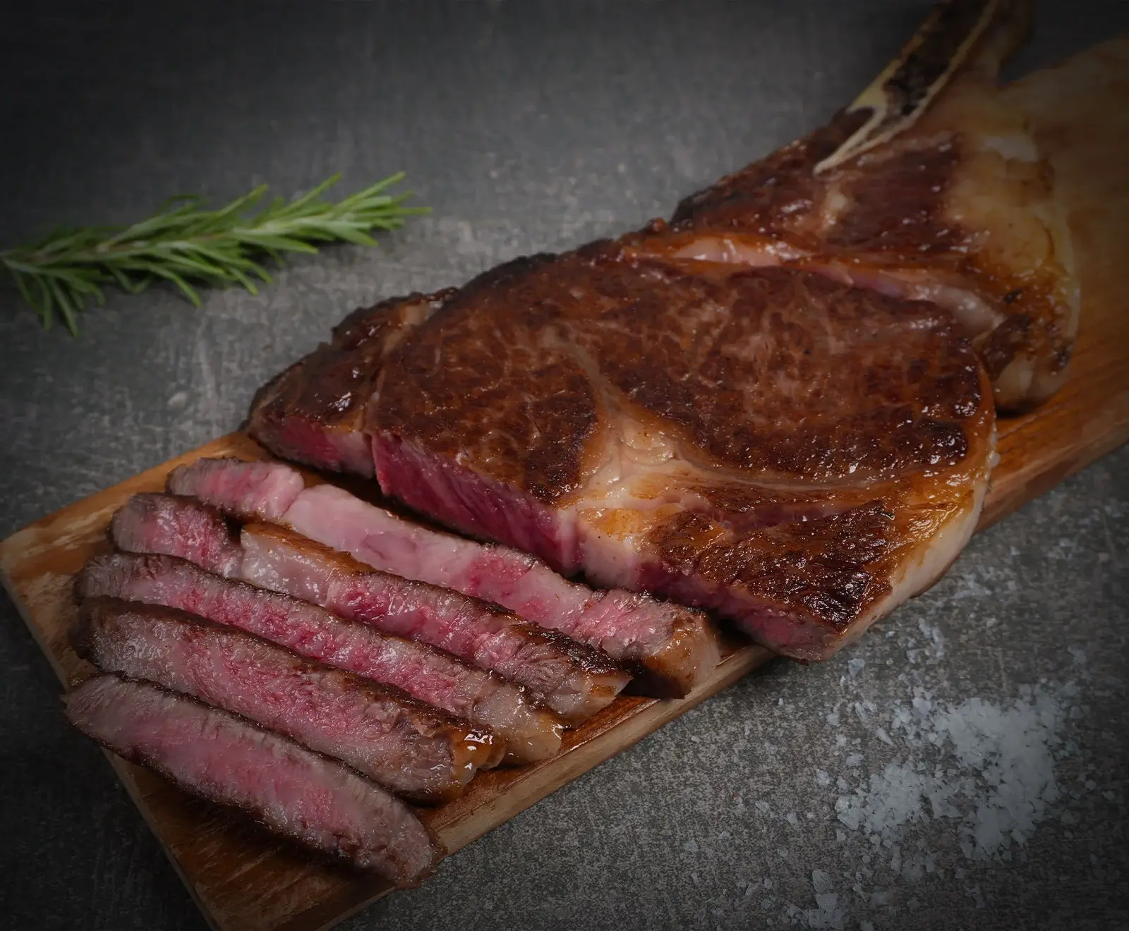 Ein perfekt gebratenes, teilweise geschnittenes Tomahawk Steak auf einem Holzbrett, mit Rosmarin und grobem Salz auf dunklem Hintergrund. Das Fleisch ist medium-rare.