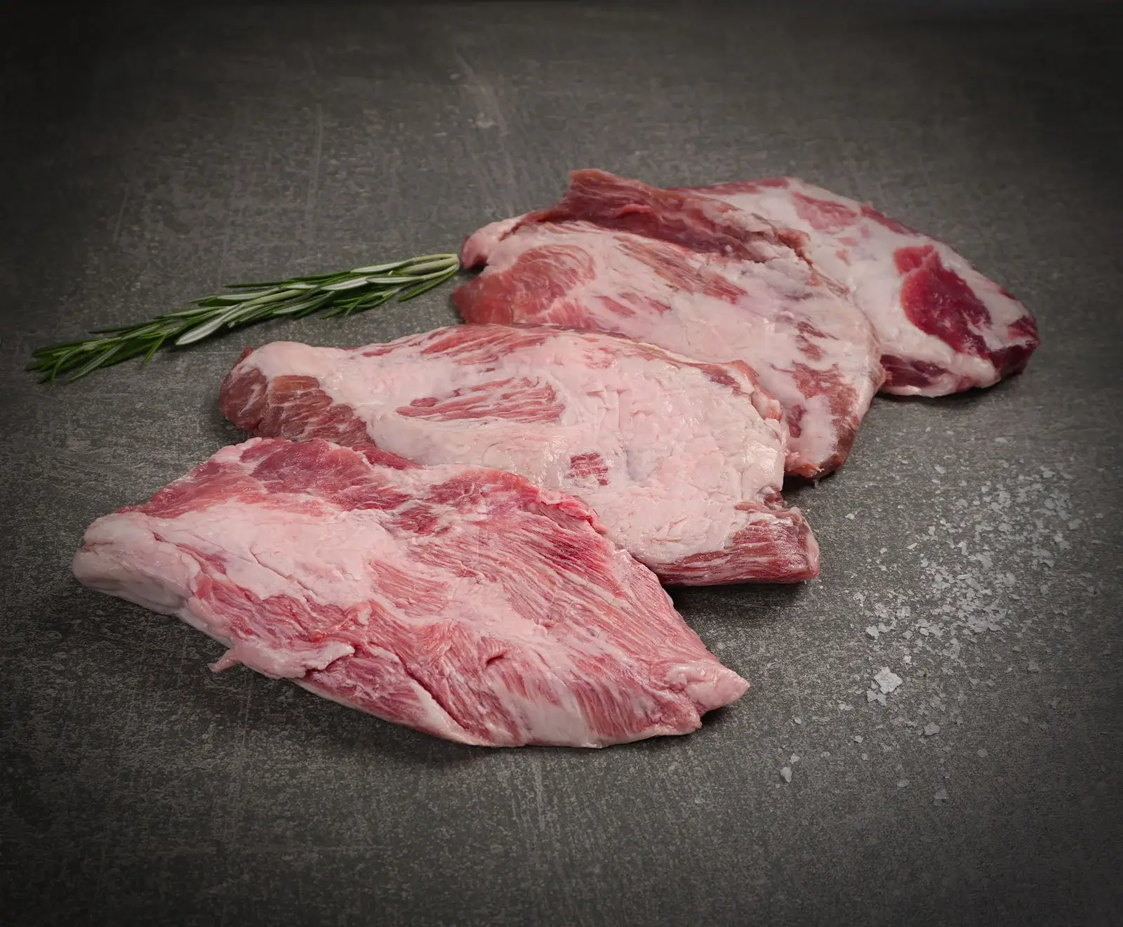 Drei Stücke rohes, marmoriertes Schweinefleisch liegen auf einer dunklen Oberfläche, begleitet von einem Rosmarinzweig und groben Salzkristallen.