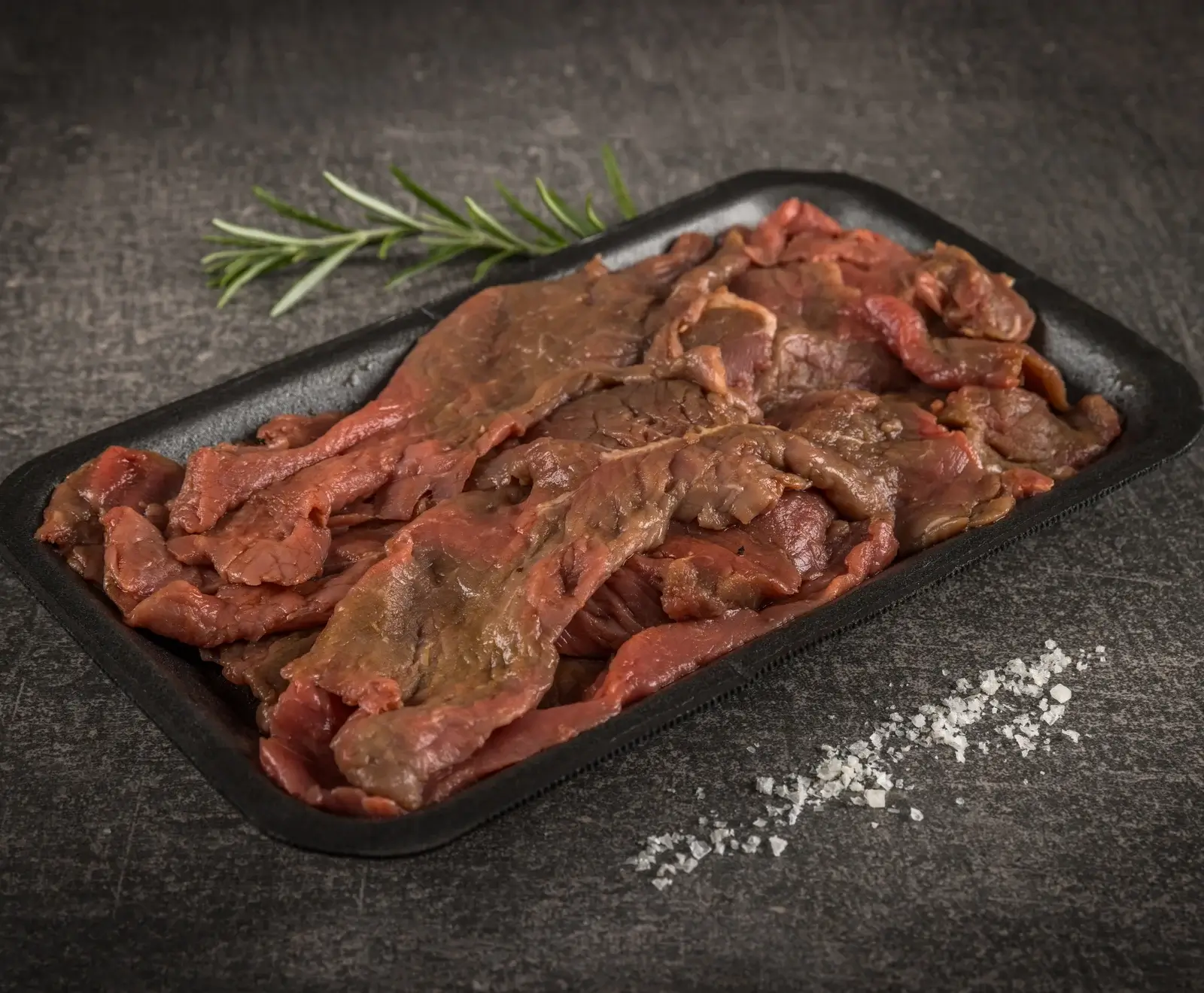 Eine schwarze Schale gefüllt mit rohen, dünn geschnittenen Rindfleischstreifen auf dunklem Untergrund. Daneben liegen ein Rosmarinzweig und grobe Salzkristalle.