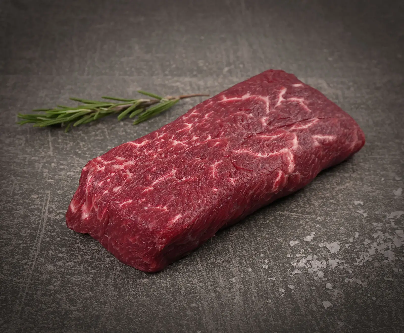 Ein rohes, marmoriertes Rindersteak liegt auf einer dunklen Steinplatte. Daneben befindet sich ein Rosmarinzweig und einige grobe Salzkristalle sind verstreut.