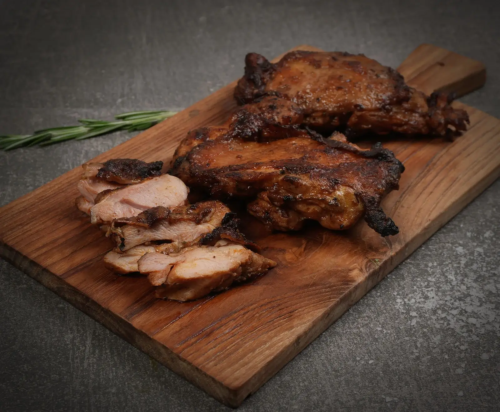 Drei goldbraun gebratene Hähnchenschenkel ohne Knochen, einer davon in Scheiben geschnitten, liegen auf einem rustikalen Holzschneidebrett. Ein Rosmarinzweig ist im dunklen Hintergrund zu sehen.
