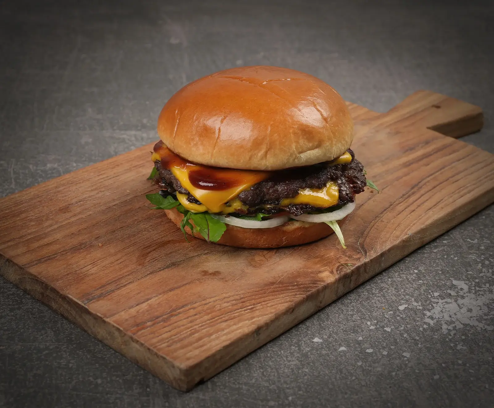 Ein appetitlicher Doppel-Cheeseburger mit glänzendem Brioche-Brötchen, zwei Patties, geschmolzenem Käse, dunkler Sauce, Rucola und Zwiebelringen auf einem Holzschneidebrett vor dunklem Hintergrund.