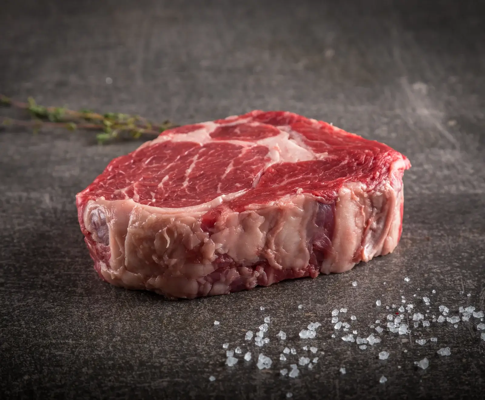Ein Stück rohes Rindfleisch auf einer dunklen Oberfläche mit Salzkristallen bestreut und einem Rosmarinzweig daneben.