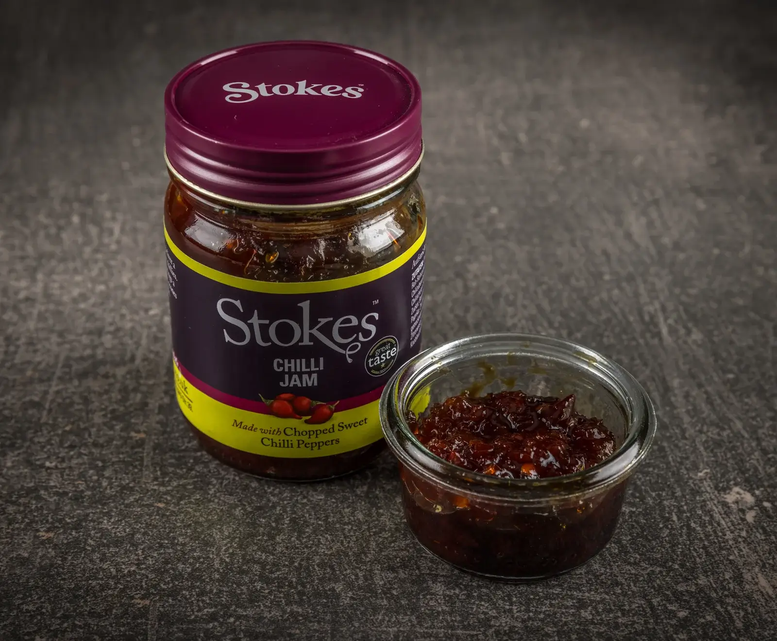 Ein Glas Stokes Chilli Jam mit lila Deckel und Etikett steht neben einer kleinen Glasschale, die mit dem dunkelroten, stückigen Chili-Marmelade gefüllt ist, auf einem dunklen, strukturierten Untergrund.