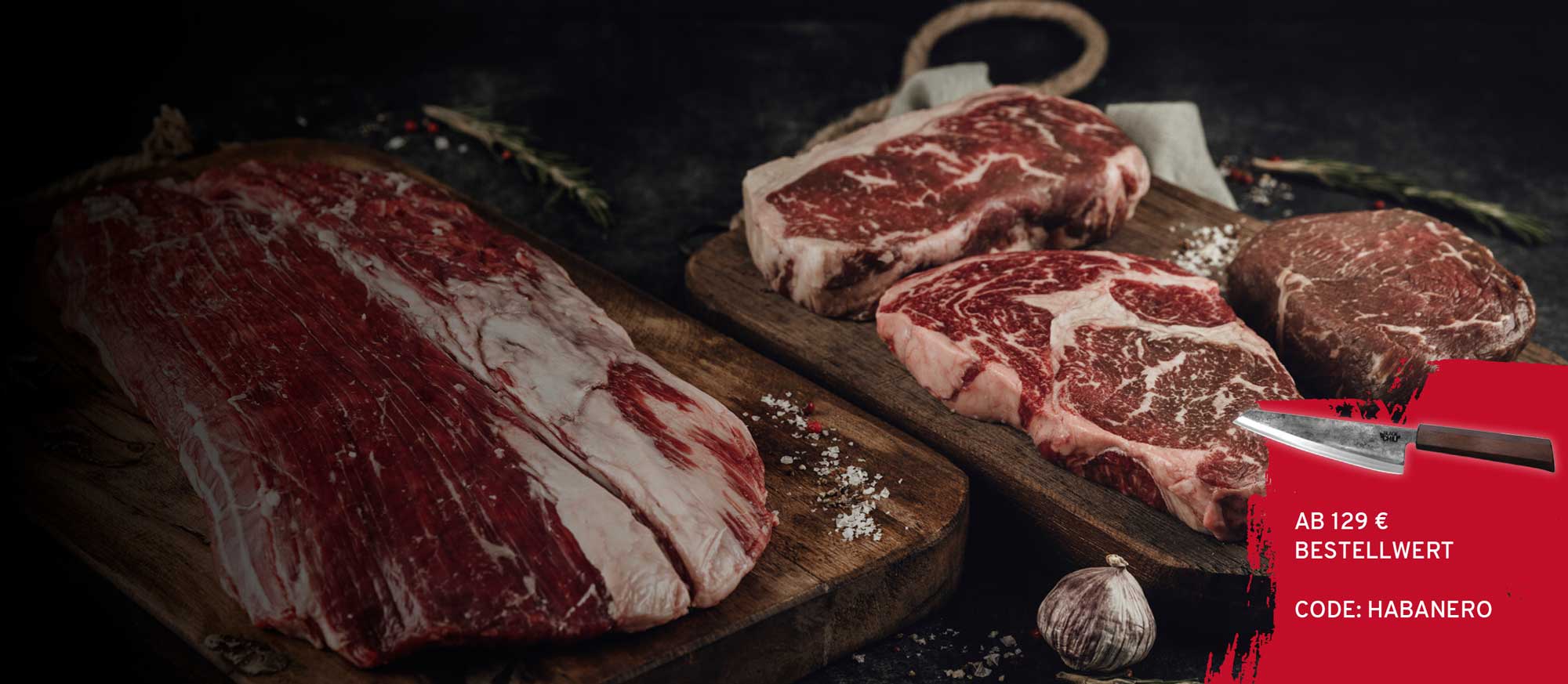 Verschiedene rohe Fleischstücke, darunter ein großes Flanksteak und drei Steaks, liegen auf Holzbrettern, bestreut mit Salz und Pfeffer. Im Vordergrund ein Knoblauch und ein rotes Banner mit Messer und Text 'AB 129 € BESTELLWERT CODE: HABANERO'.