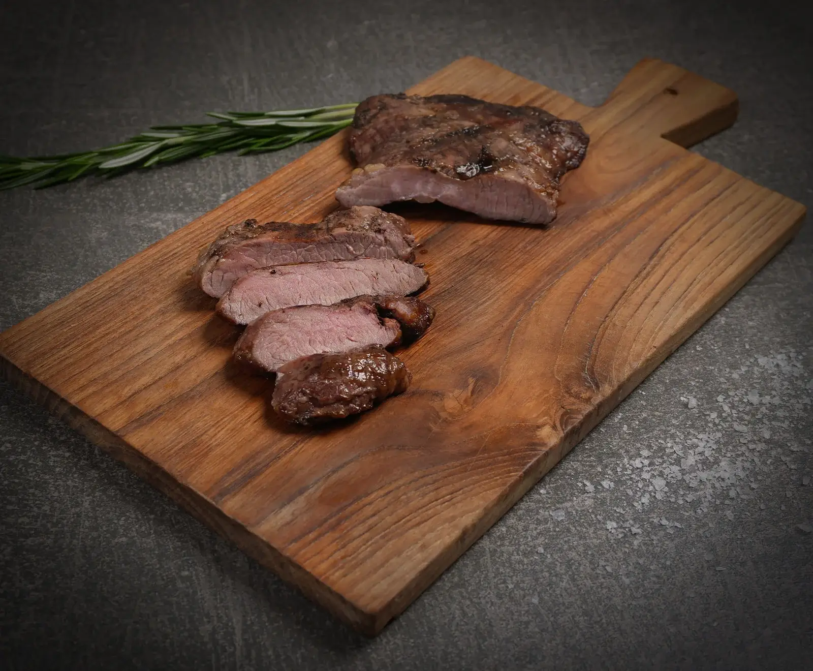 Ein aufgeschnittenes, rosa gebratenes Stück Fleisch liegt auf einem rustikalen Holzschneidebrett. Daneben befindet sich ein Rosmarinzweig auf dunklem Untergrund.