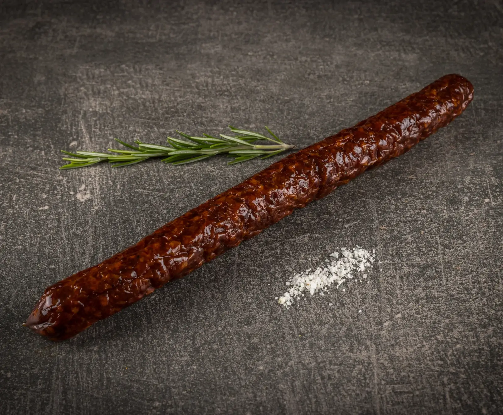Eine lange, dunkelbraune Rohwurst liegt diagonal auf einer dunklen, strukturierten Oberfläche, begleitet von einem Rosmarinzweig und einer Prise grobem Salz.