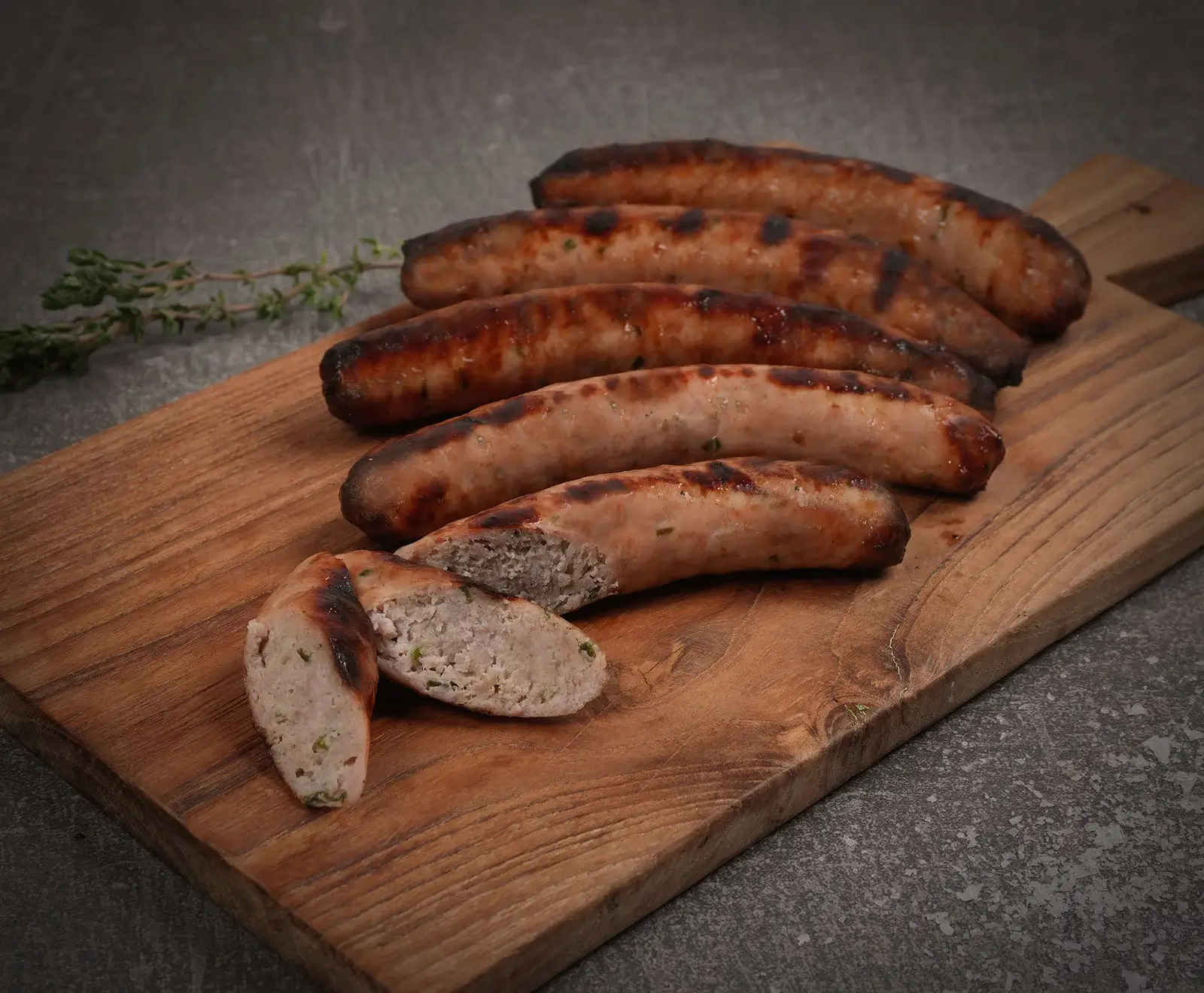 Fünf goldbraun gegrillte Würste und eine halbierte Wurst mit sichtbarer Füllung liegen auf einem rustikalen Holzbrett. Ein frischer Thymianzweig ist links zu sehen.