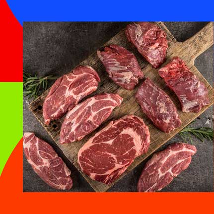 Verschiedene rohe Fleischstücke, darunter Steaks und andere Schnitte, liegen auf einem rustikalen Holzschneidebrett. Einige Stücke zeigen deutliche Marmorierung. Ein Rosmarinzweig ist ebenfalls zu sehen.