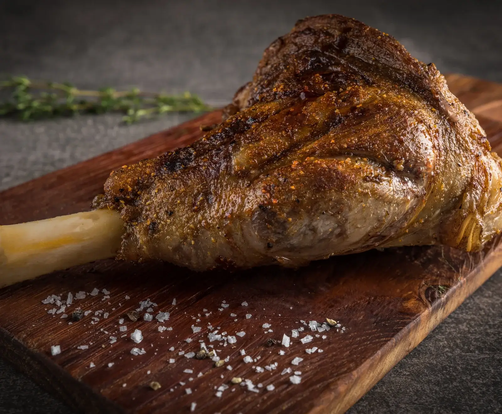 Eine goldbraun gebratene Lammkeule mit knuspriger Haut und sichtbarem Knochen liegt auf einem dunklen Holzbrett. Grobes Salz und Pfeffer sind auf dem Brett verstreut, ein Thymianzweig im unscharfen Hintergrund.