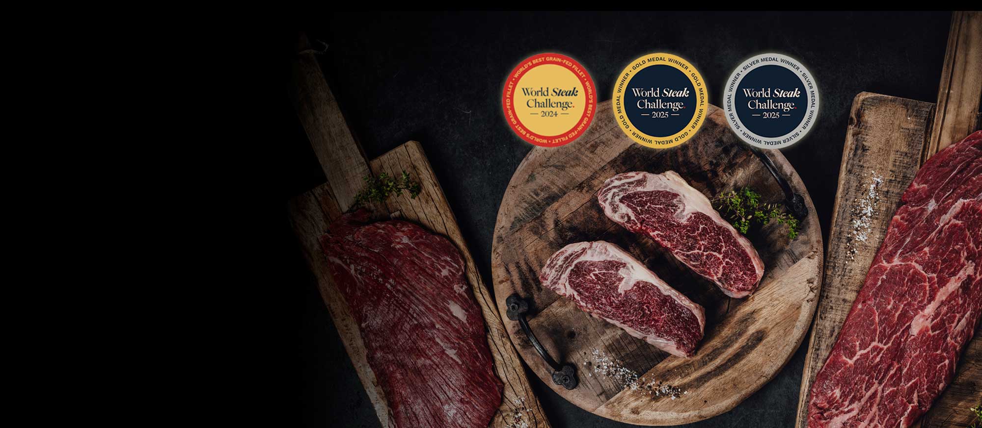Drei rohe Rindfleischstücke mit Marmorierung auf Holzschnitten, darunter zwei Ribeye-Steaks. Darüber drei World Steak Challenge Medaillen (2024: Bestes Grain-Fed Filet, 2025: Gold, Silber). Dunkler Hintergrund.