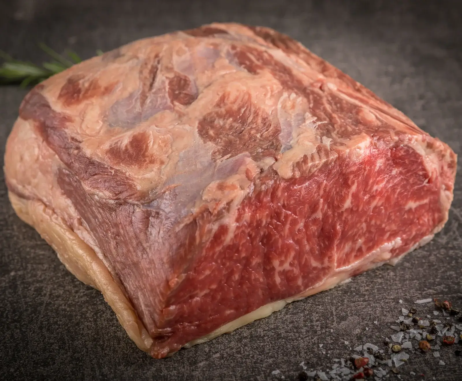 Ein großes Stück rohes Rindfleisch mit deutlicher Marmorierung und Fettrand liegt auf einer dunklen Oberfläche. Im Hintergrund Rosmarin, im Vordergrund Salz und Pfeffer.