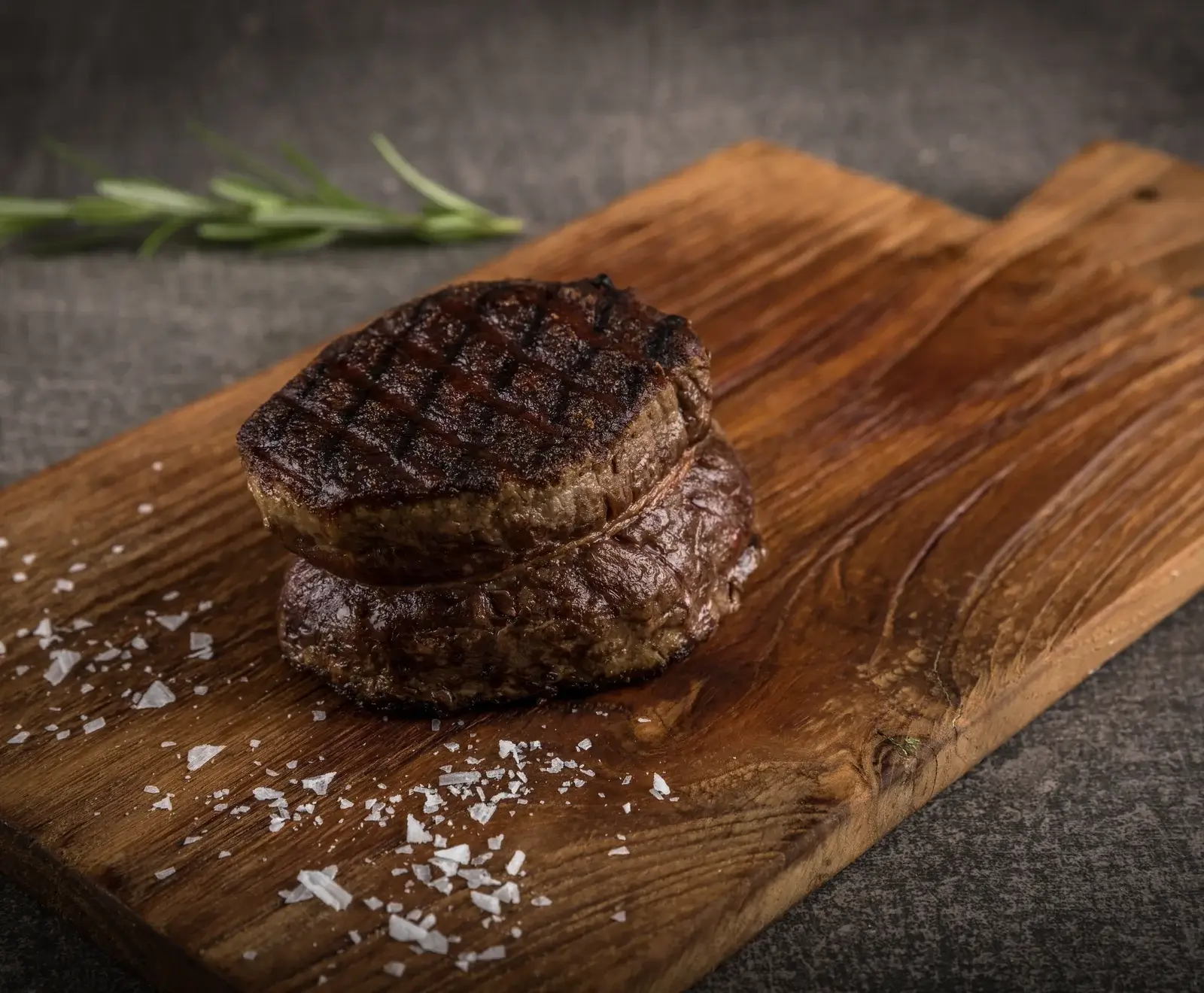 Zwei saftige, gegrillte Steaks mit Grillstreifen, übereinander auf einem rustikalen Holzbrett, bestreut mit grobem Meersalz. Im unscharfen Hintergrund liegt ein Rosmarinzweig.