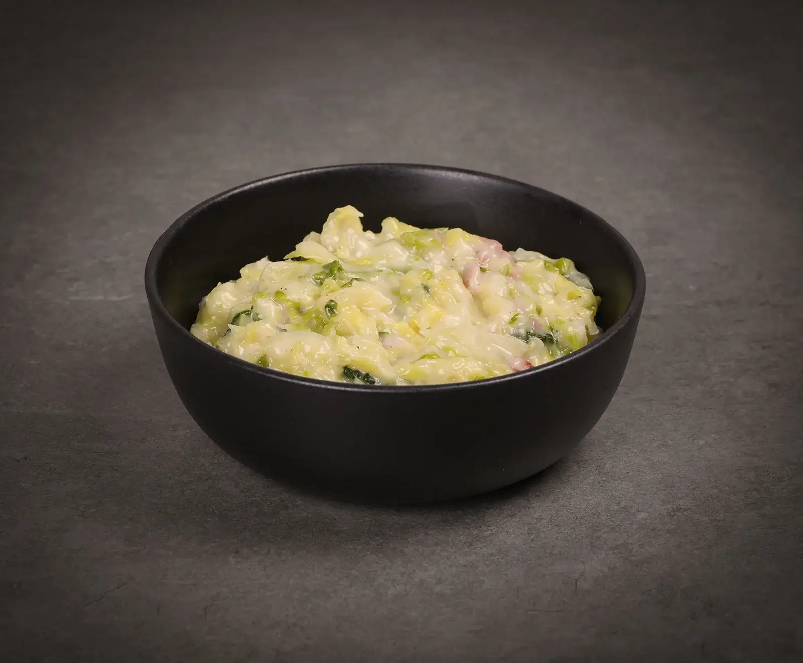 Eine schwarze Schale gefüllt mit cremigem Kartoffel-Lauch-Eintopf, angereichert mit kleinen Speckwürfeln, auf einem dunklen, strukturierten Hintergrund.