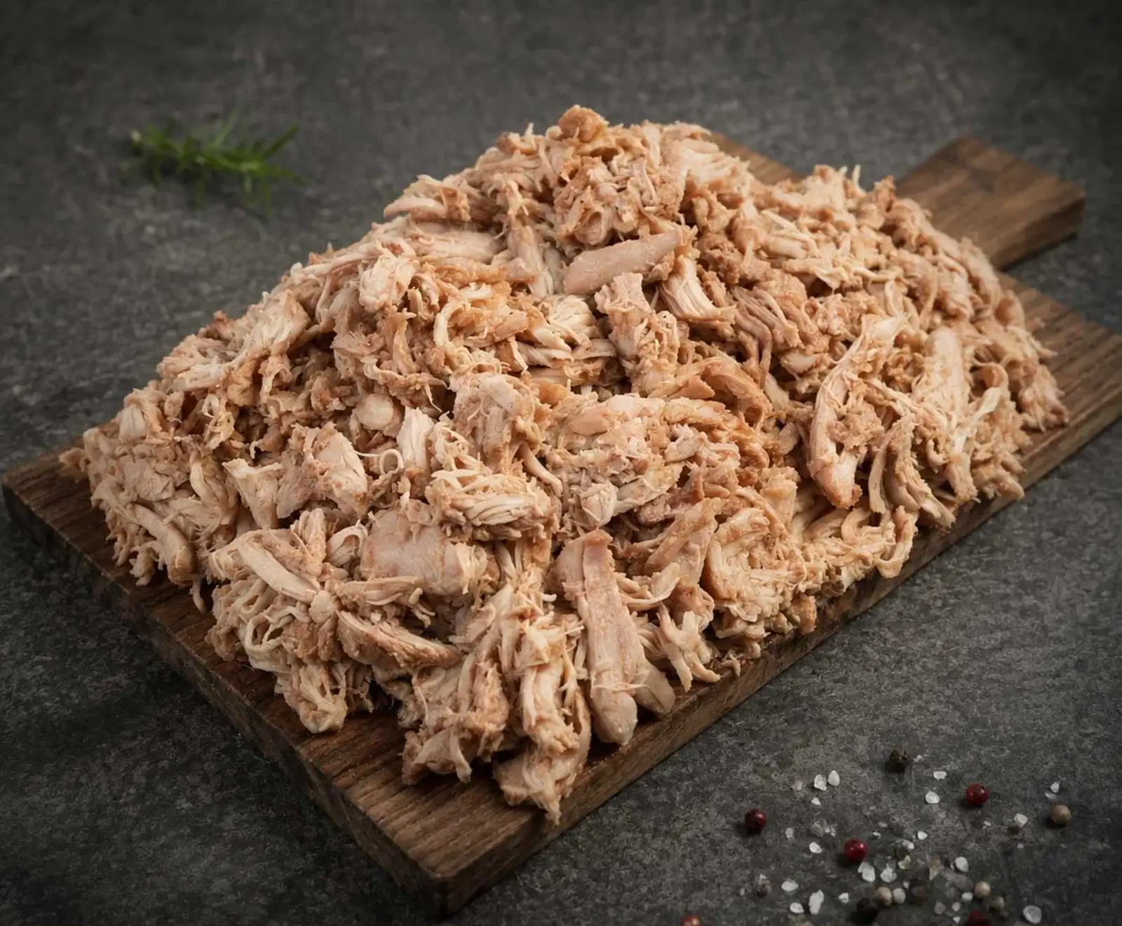 Ein großer Haufen zerkleinertes, gekochtes Hähnchenfleisch (Pulled Chicken) auf einem rustikalen Holzbrett. Im Hintergrund ein Rosmarinzweig, im Vordergrund verstreute Pfefferkörner und grobes Salz.