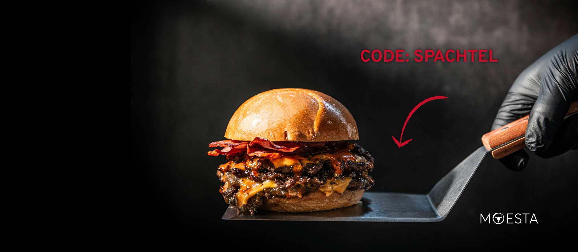 Ein saftiger Burger mit zwei Patties, geschmolzenem Käse und knusprigem Speck auf einem Brötchen, der auf einem Metallspatel liegt. Eine behandschuhte Hand hält den Spatel. Oben rechts steht "CODE: SPACHTEL" mit einem roten Pfeil, der auf den Spatel ze